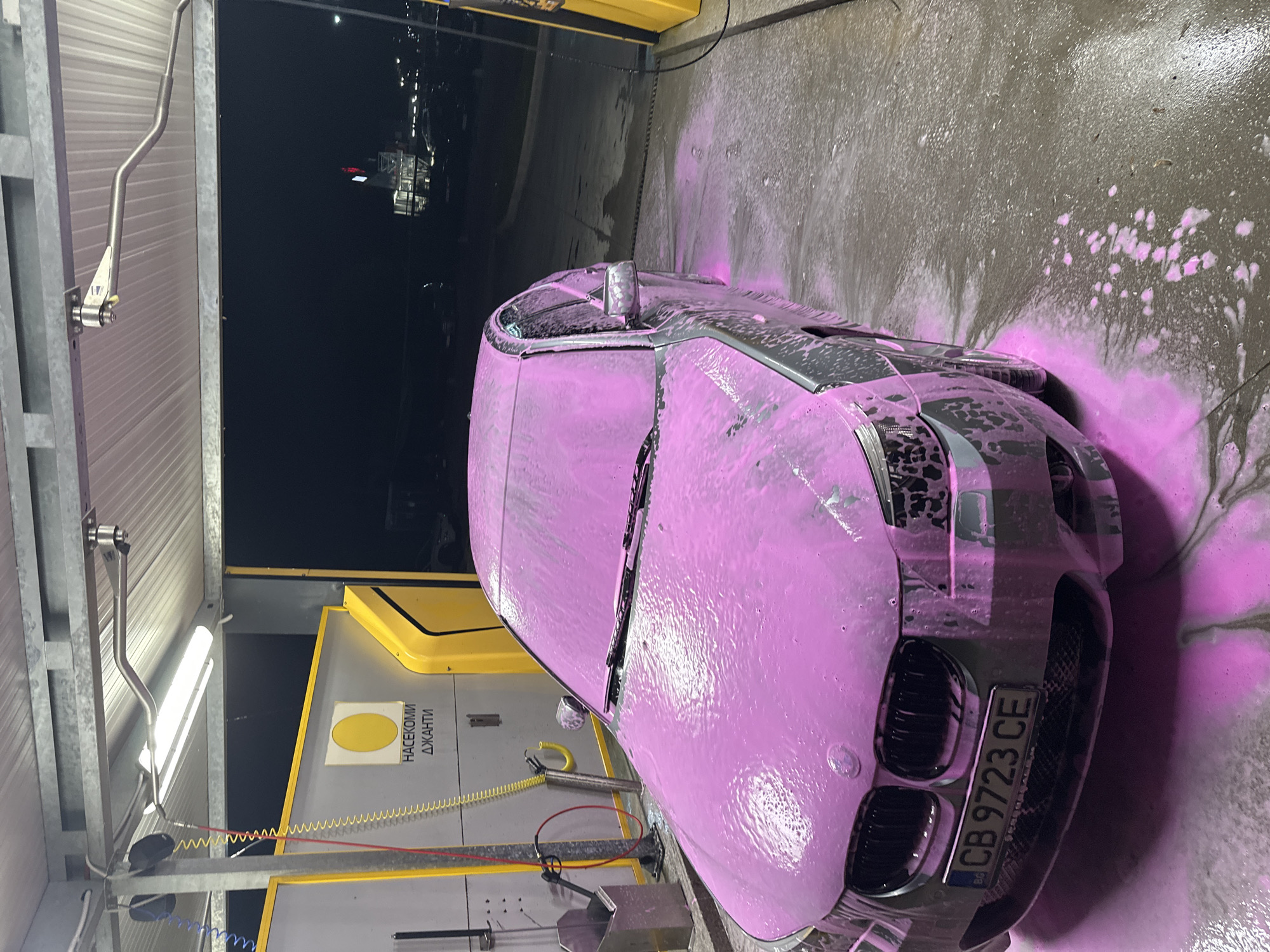 A car is covered in pink foam at a car wash. The license plate reads "CB 9723 CE." The car wash is located in Plovdiv, Bulgaria, indicated by the license plate. The background is dark, suggesting it is nighttime. The car is parked in a wash bay with cleaning equipment visible. A sign in Bulgarian is on the yellow structure.