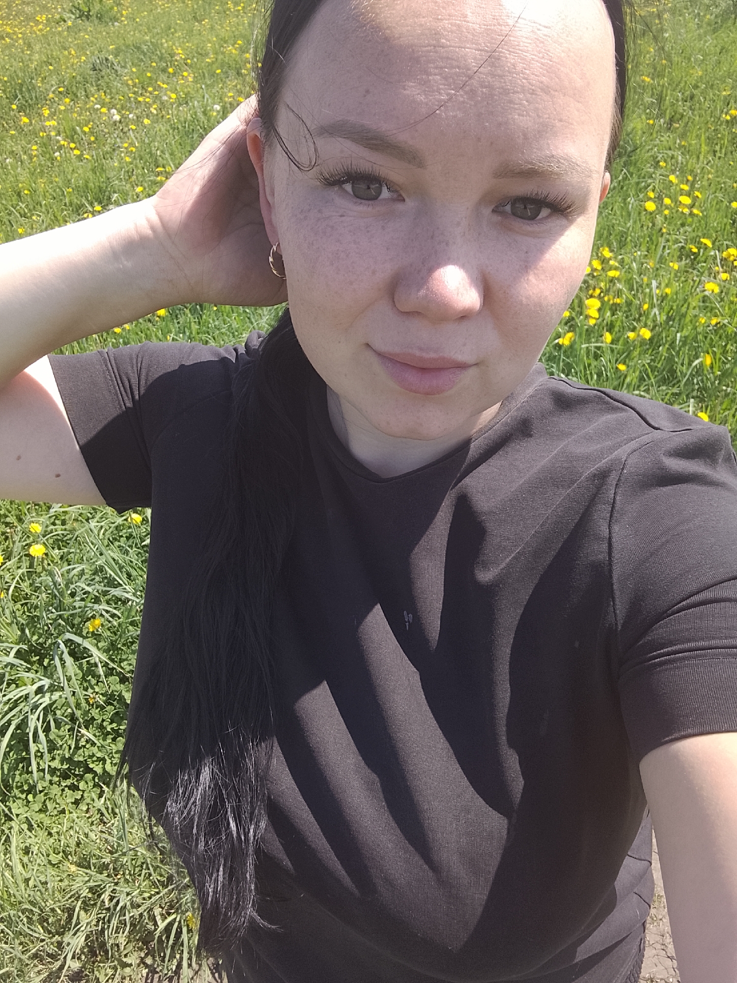 A fair-skinned woman with freckles is taking a selfie in an area of grass and yellow wildflowers. She is wearing a dark T-shirt and has dark hair pulled back. She is holding her hand near her ear, and part of her hair is visible in front of her chest. The location cannot be determined as Кипчак, Russia from visual information.