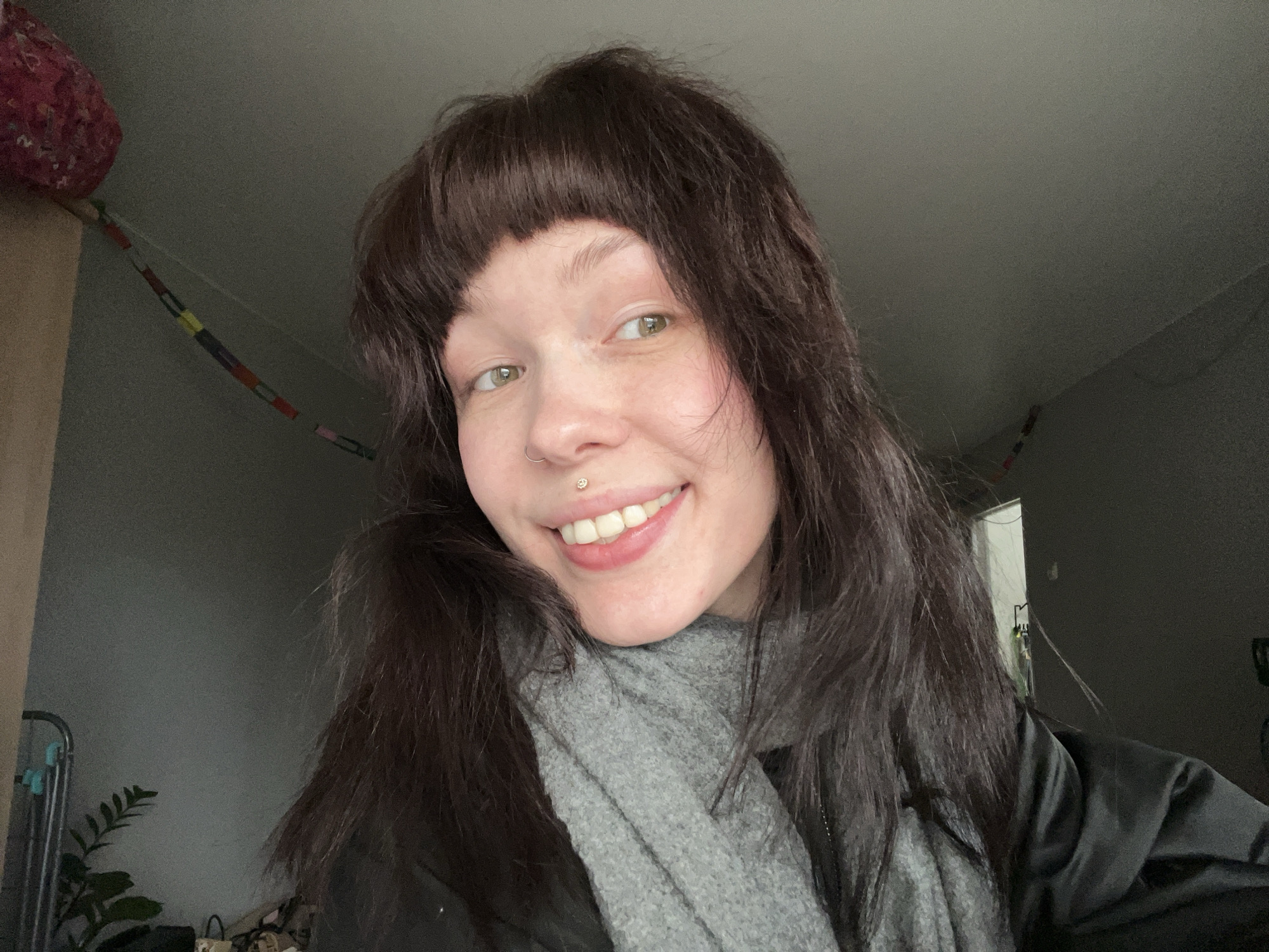A selfie of a woman with light skin, brown hair with short bangs, and green eyes. She is smiling and wearing a gray scarf and a black jacket. A lip piercing with a stud is visible. The background shows a dimly lit room with a window. There is also a plant, a garment rack, and a handmade paper chain. A red object is on the top left. The image appears to be taken indoors. There is no evidence of the location being Chaikovsky, Russia.
