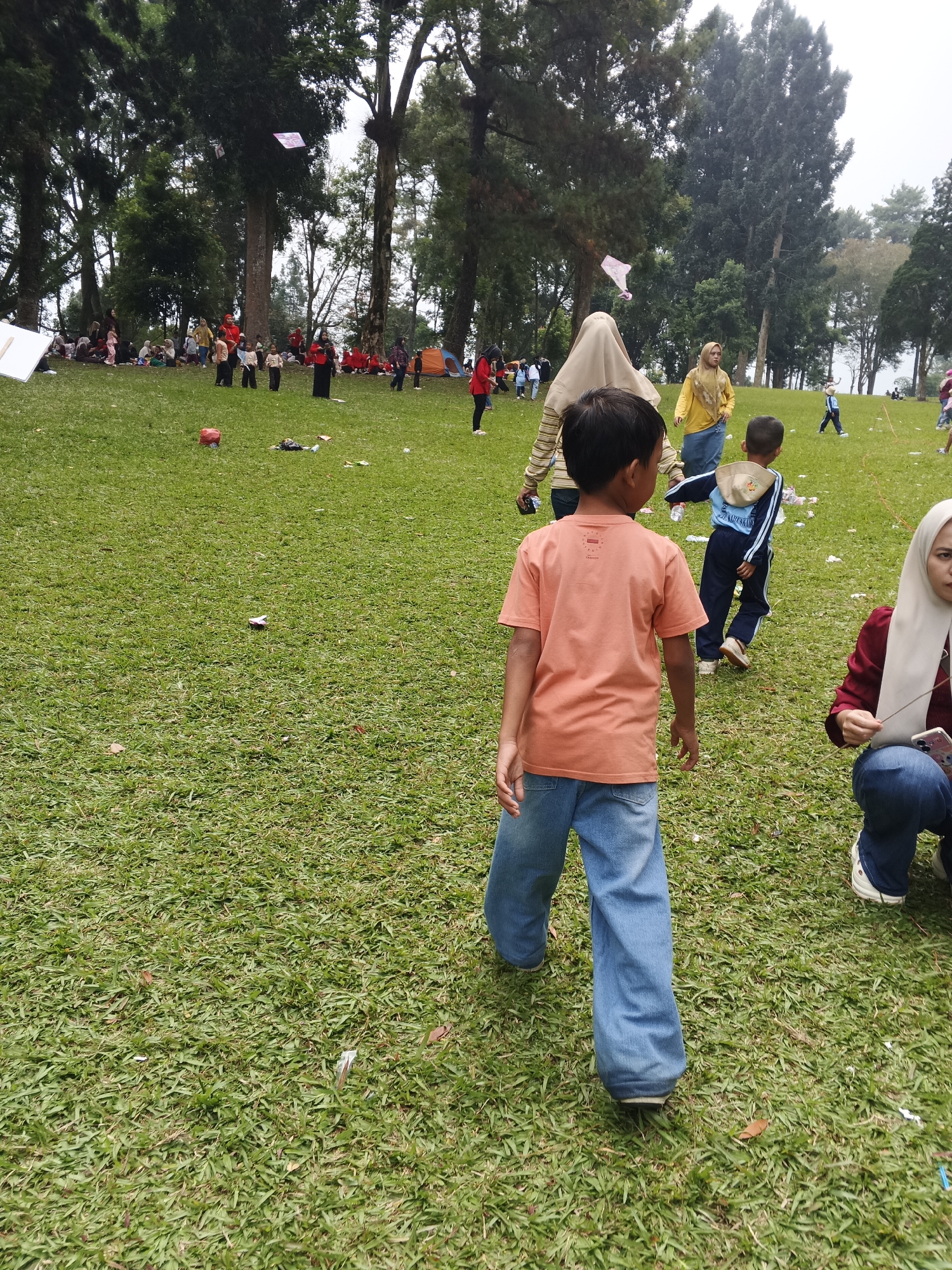 A group of people, including children and adults, are gathered on a grassy field in a park, possibly in the city of Perbawati, Indonesia. The scene is set with large trees providing shade in the background. Some individuals are sitting on the ground, while others are standing or walking. One child in the foreground is wearing an orange t-shirt and blue jeans, facing away from the camera and walking towards the right of the frame. To the right of this child, another child is wearing a blue tracksuit and a hat. In the middle ground, several people are seated on the grass, some wearing hijabs. Further in the background, there are more people, tents, and kites flying in the air. The overall atmosphere appears to be a casual outdoor gathering or event.