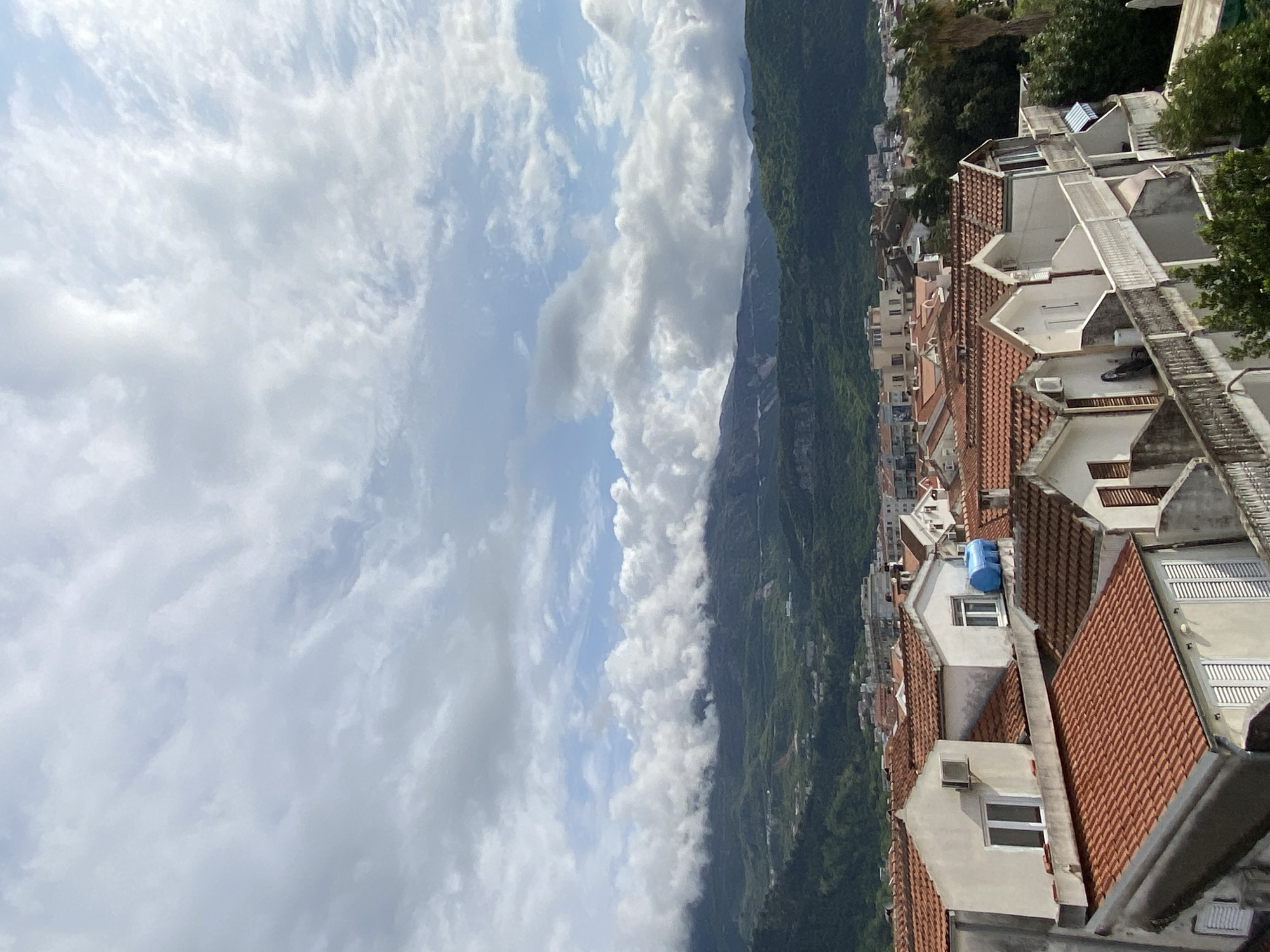 The image captures a panoramic daytime view of a town situated at the base of lush green mountains, under a dynamic, partly cloudy sky. The perspective is from an elevated vantage point, looking down across a dense cluster of buildings.
In the foreground and mid-ground, numerous residential structures with characteristic red terracotta tiled roofs dominate the scene. Their facades are predominantly white or light-colored, and many feature small balconies and rectangular windows. Air conditioning units are visible on the exteriors of some buildings, and one roof distinctly displays a bright blue, cylindrical water tank or similar object. The architectural style suggests a Mediterranean or European coastal town setting.
Beyond the urban sprawl, a sprawling range of verdant, tree-covered mountains extends into the distance. These mountains are partly enveloped by clouds, creating a hazy, atmospheric effect. The sky above is a patchwork of light blue areas and scattered white and gray clouds, indicating fair weather. No people or specific activities are visible, presenting a tranquil and scenic cityscape. The overall impression aligns with coastal regions known for their mountainous backdrops and traditional architecture, such as Budva, Montenegro. There is no visible text in the image.