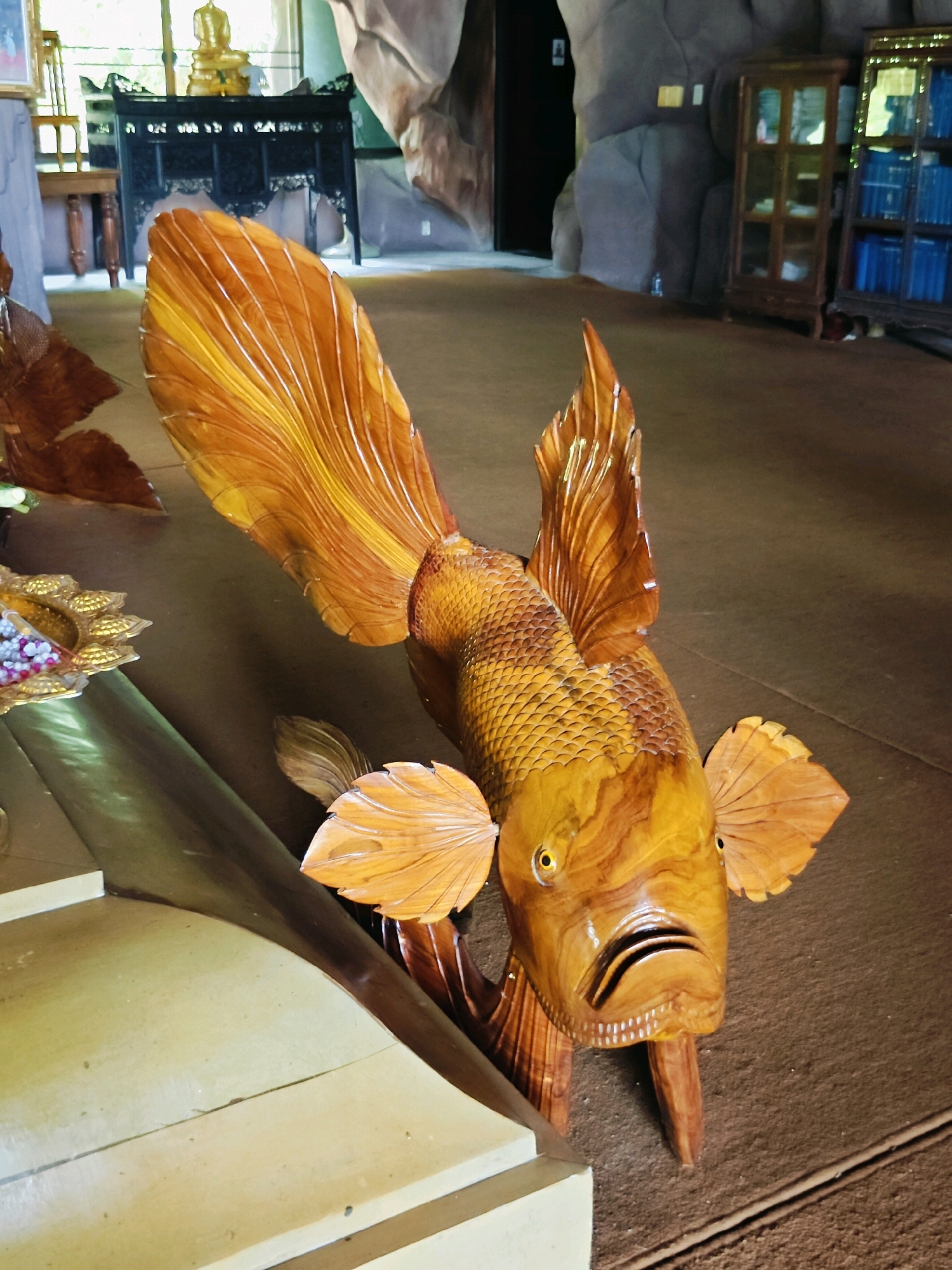 The image shows a wooden sculpture of a large fish with exaggerated fins and detailed scales, displayed indoors on a carpeted floor. The fish is carved from a light-colored wood with darker grain patterns, and it has a wide, open mouth with visible teeth. Its eyes are round and yellow. In the background, there are elements suggestive of an indoor environment, possibly a temple or a decorative space. To the left, a portion of a light-colored structure with a curved edge is visible, and beyond that, a dark wooden cabinet with carvings and a golden Buddha statue are partially seen. To the right, a tall, dark wooden cabinet with glass doors containing rows of blue items is visible, and behind it, a rocky, textured wall or structure. The overall setting appears to be a room with various decorative objects, suggesting it might be a place of cultural or religious significance. The location is Hantra Subdistrict, Thailand.