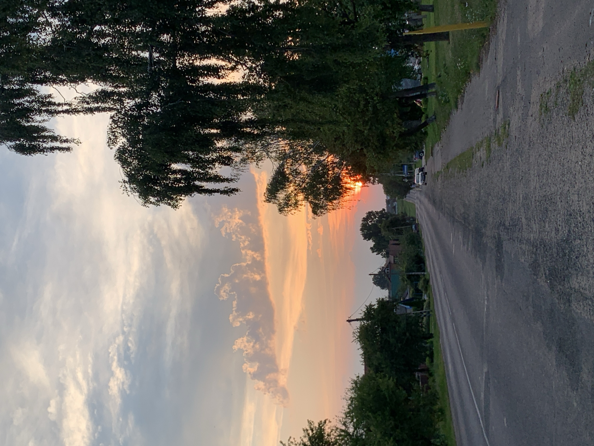 A rural road in Kapyĺ, Belarus, stretches towards a sunset. The sky is a gradient of pinks, oranges, and blues, with scattered clouds. Lush green trees line the right side of the road, their branches hanging low. On the left, houses and more trees are visible in the distance. A paved road with a white line down the center dominates the foreground. Two cars are parked on the right side of the road. The scene is tranquil and evokes a sense of the end of a day.