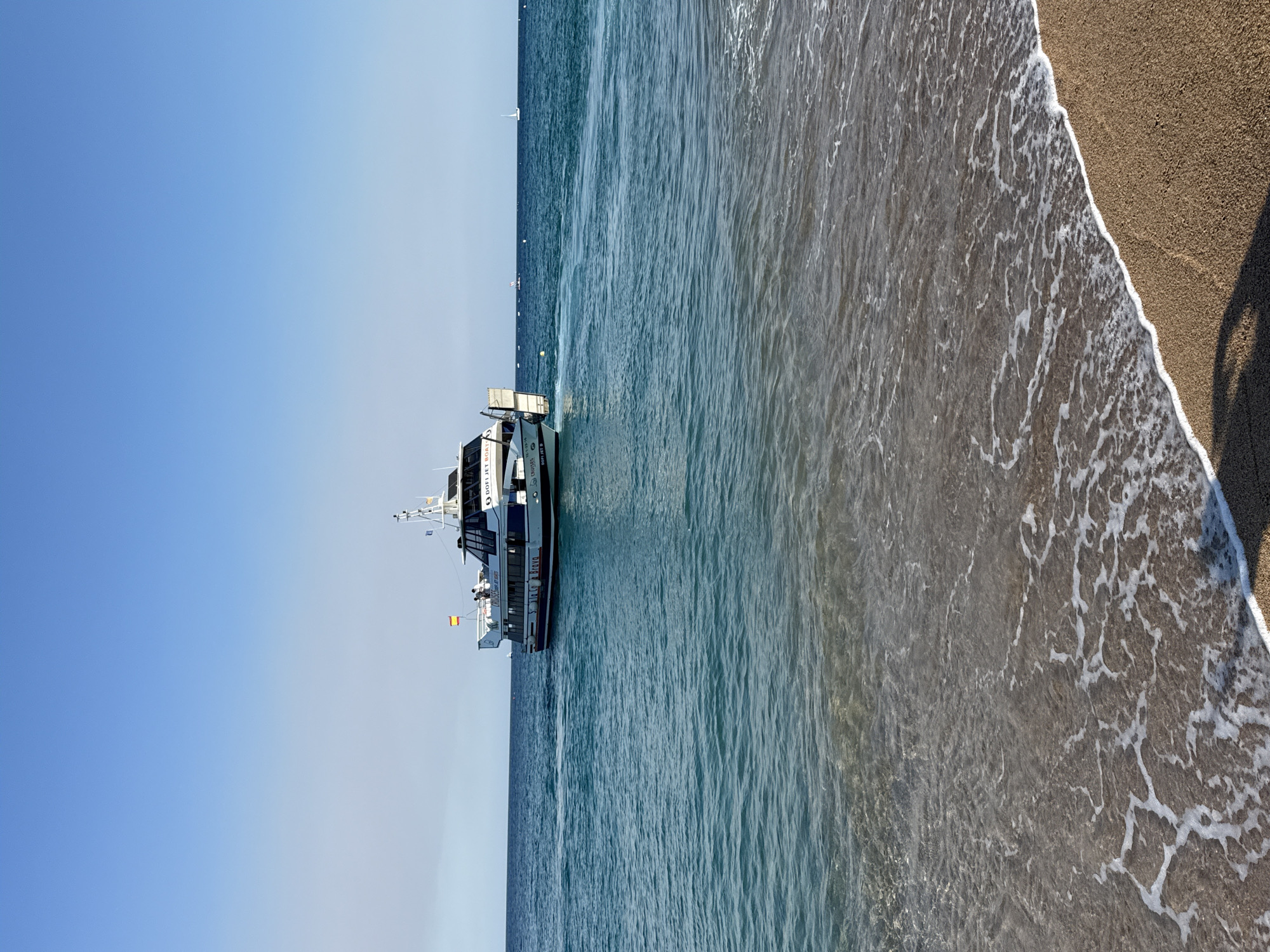 In Blanes, Spain, a white and blue passenger boat with the name "SILA BRAVO" on its side is sailing on the azure waters of the Mediterranean Sea. The boat, which appears to be a ferry or tour boat, has several people on its upper deck. A Spanish flag is visible at the stern of the boat. The water is calm, with small waves lapping at the sandy shore in the foreground. In the distance, two sailboats can be seen on the horizon under a clear blue sky. The scene captures a peaceful day at the coast of Blanes.