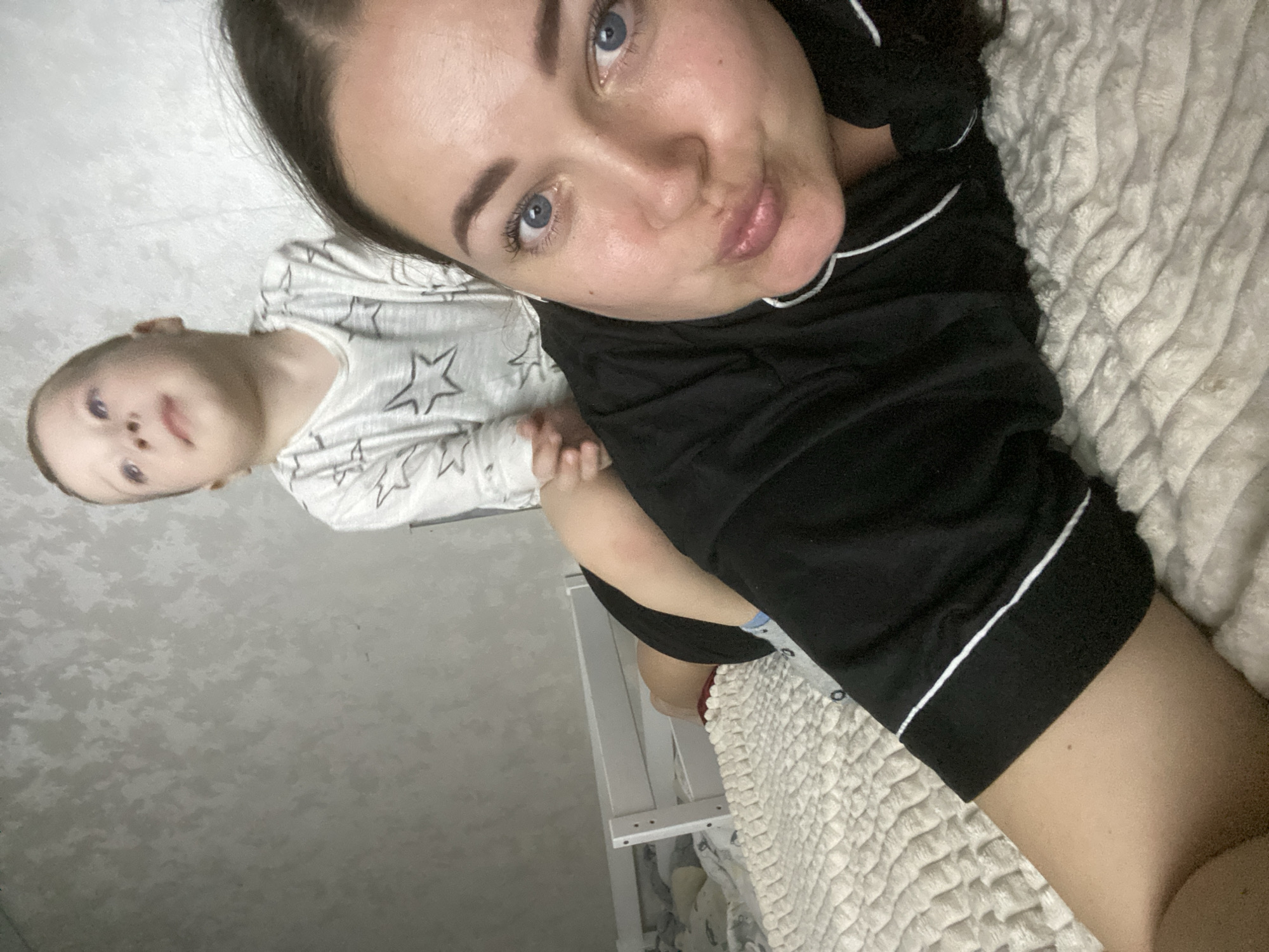 A woman and a toddler are lying on a bed, possibly in a room. The woman is in the foreground, wearing black pajamas with white trim, and her face is close to the camera. The toddler is wearing a light-colored top with star patterns. The bed has a textured white cover. Part of a white crib is visible. The location in the city of Sochi, Russia is unknown.