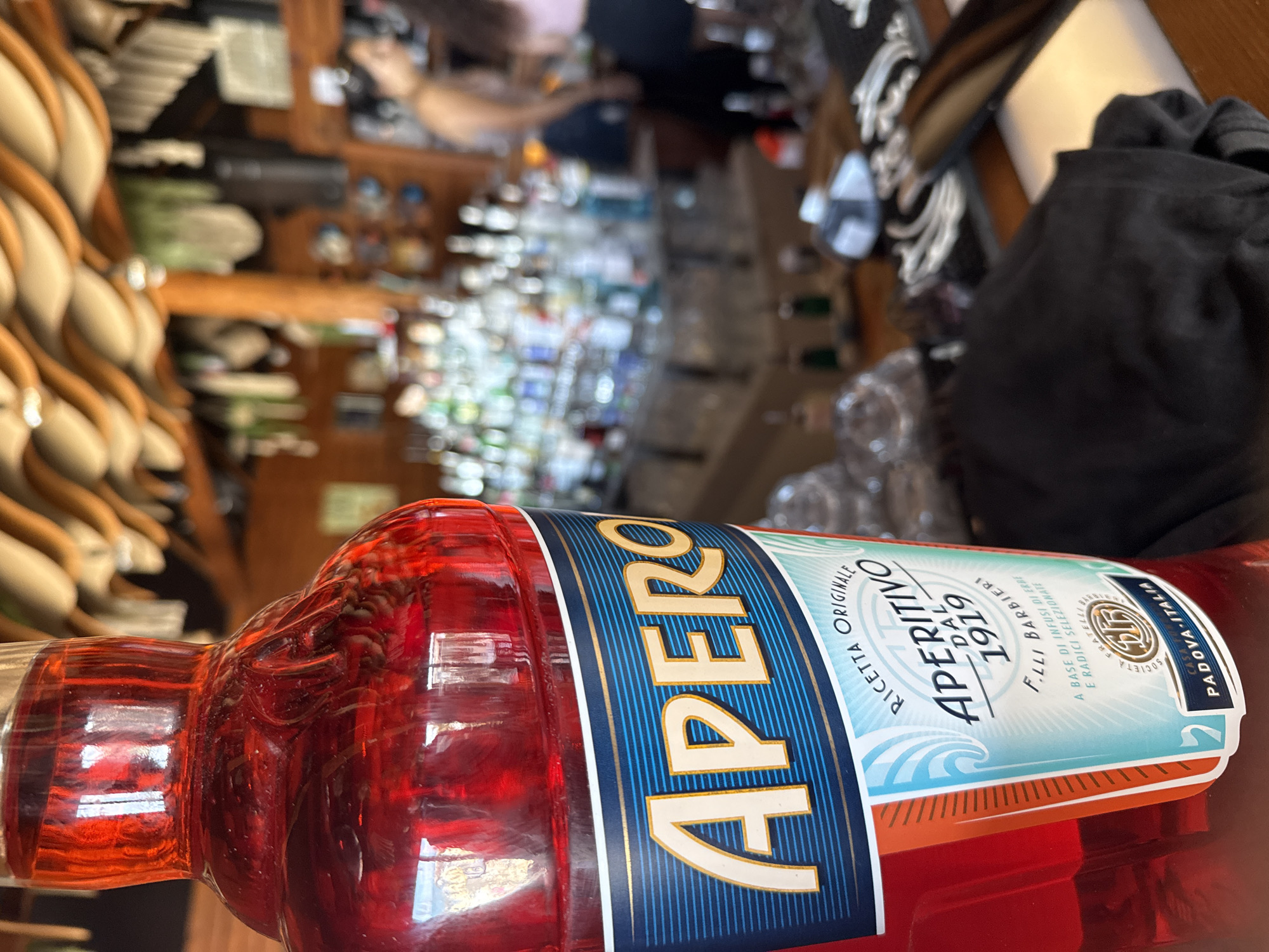 A close-up shot features a bottle of Aperol, a red liqueur, prominently displayed in the foreground. The bottle's label, with bold "APEROL" lettering, is clearly visible. Behind the bottle, a blurred background reveals a bar area. Shelves filled with various bottles of alcohol line the back wall. Two individuals, out of focus, are visible in the background. One person appears to be a woman working behind the bar, while another person stands nearby. The overall setting suggests a bar or restaurant environment. The specific location in the city of Pomorie, Bulgaria, is not discernible from the image.