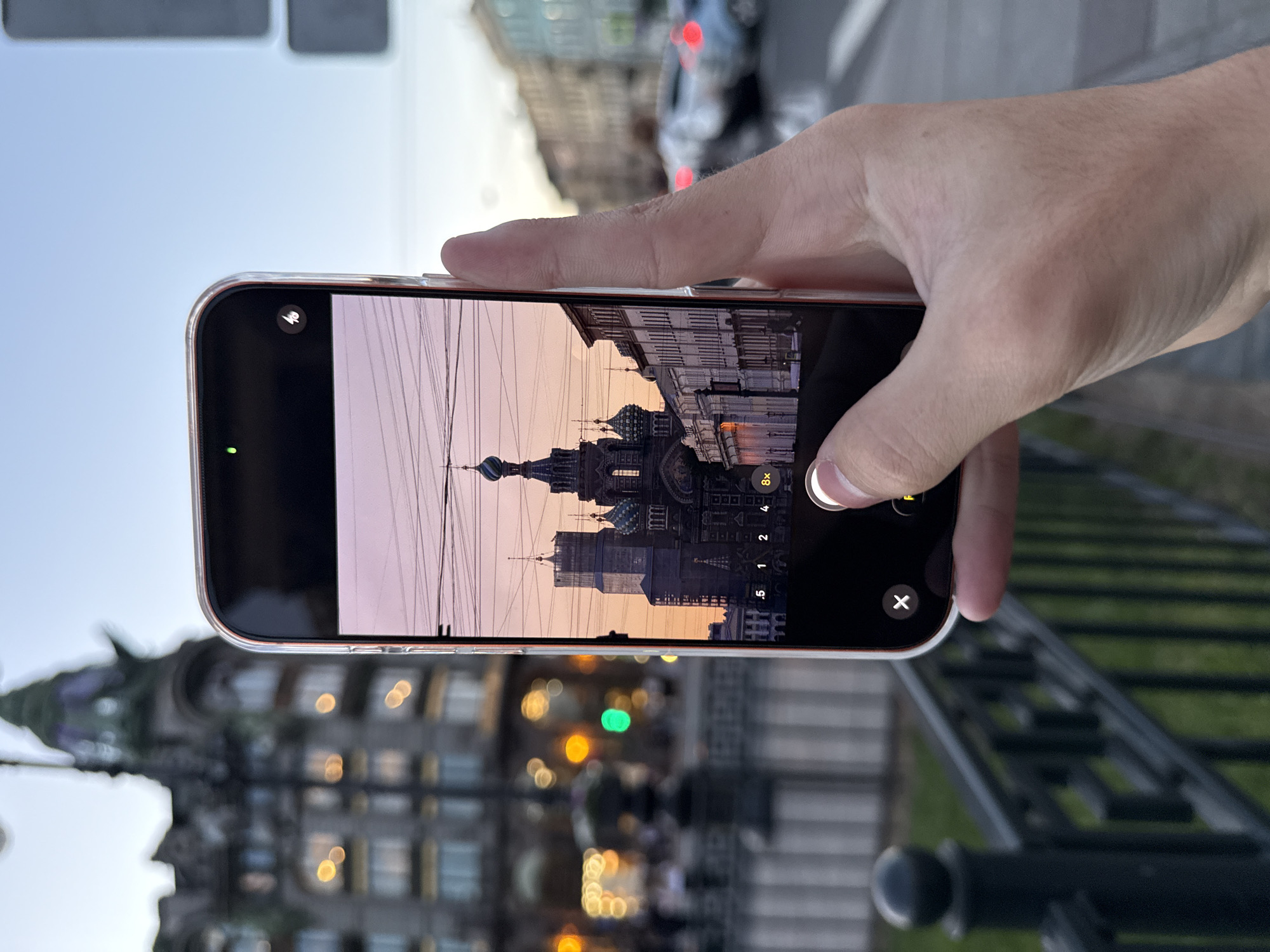 A close-up shot captures a person holding a smartphone, framing a view of the Church of the Savior on Spilled Blood in Saint Petersburg, Russia. The church's distinctive onion domes are visible against a twilight sky with soft pink and orange hues. Power lines crisscross the sky, adding to the urban landscape. In the foreground, a dark metal fence with rounded posts is partially visible. The phone's camera interface shows zoom options, including "8x," and a button being pressed by a thumb. The background is blurred, showing the lights of buildings and moving vehicles, further indicating an urban setting.