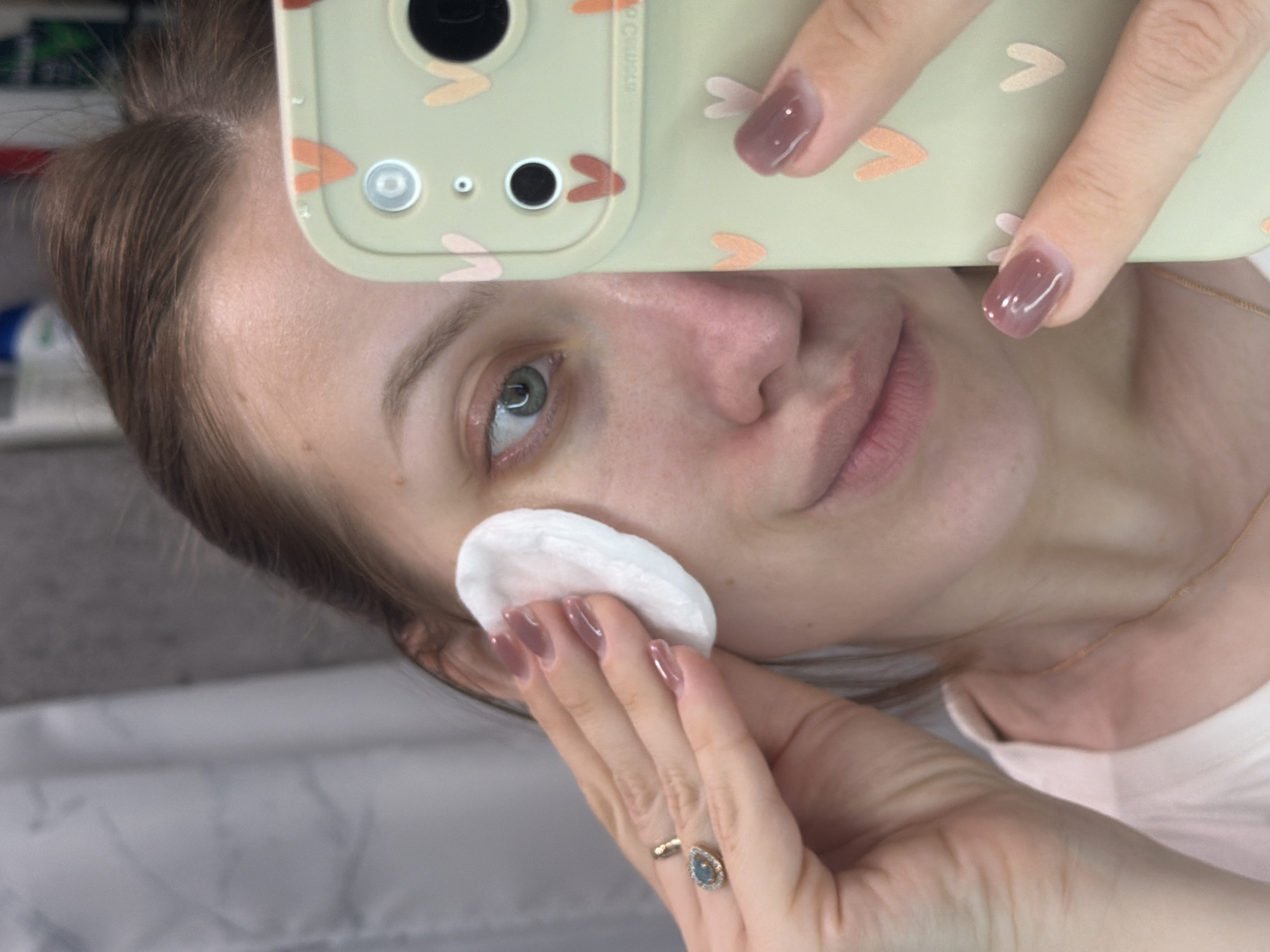 A woman is holding a cell phone with a light green case with small, stylized heart-shaped designs over her face, taking a selfie. She is holding a white cotton pad against her cheek with her other hand, which is adorned with rings and has manicured nails. She is wearing a gold necklace. The background appears to be a bathroom setting.