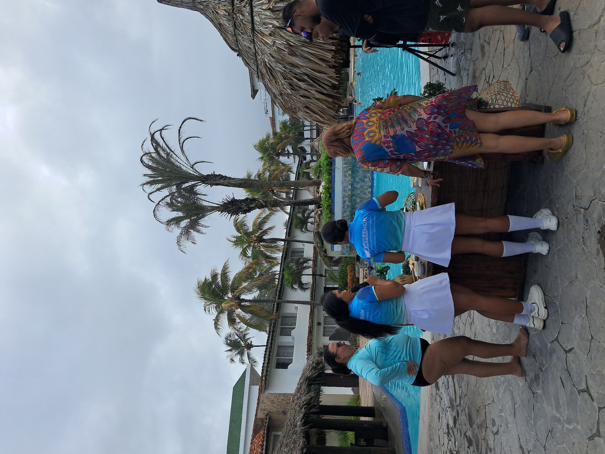 The image captures a vibrant outdoor scene at a tropical resort in El Tirano, Venezuela, under an overcast sky. Several individuals are gathered around a swimming pool area.
In the foreground and mid-ground, three women are prominently featured near the pool's edge. Two of them wear matching light blue short-sleeved tops and white short skirts, with "ANIMACION" (Spanish for "Animation") visible on their shirts. One of these women has long dark hair and wears white sneakers and socks. The third woman, closest to the viewer, wears a light blue long-sleeved top and black bottoms, standing barefoot on the stone pavement. Her dark hair is pulled back. These three individuals appear to be staff or entertainers, possibly preparing for an activity.
To their right, a woman with reddish-brown hair is seen from behind, wearing a colorful, flowing dress with a distinctive peacock-like feather pattern. She is wearing gold flat shoes and appears to be leaning over a wooden counter or table. Further to the right and in the background, two men are standing near a thatched-roof structure. One man wears sunglasses and a black t-shirt, while the other is in a black t-shirt and patterned shorts.
The setting includes a bright blue swimming pool, surrounded by stone-paved ground. Numerous palm trees, some very tall, dot the landscape, adding to the tropical ambiance. In the background, a white multi-story building with several windows and some red-tiled roofing is visible, likely part of the resort complex. A large thatched roof provides shade over an area, possibly a bar or seating. The sky is uniformly cloudy, suggesting an overcast day rather than direct sunlight. The overall atmosphere appears relaxed and vacation-oriented.