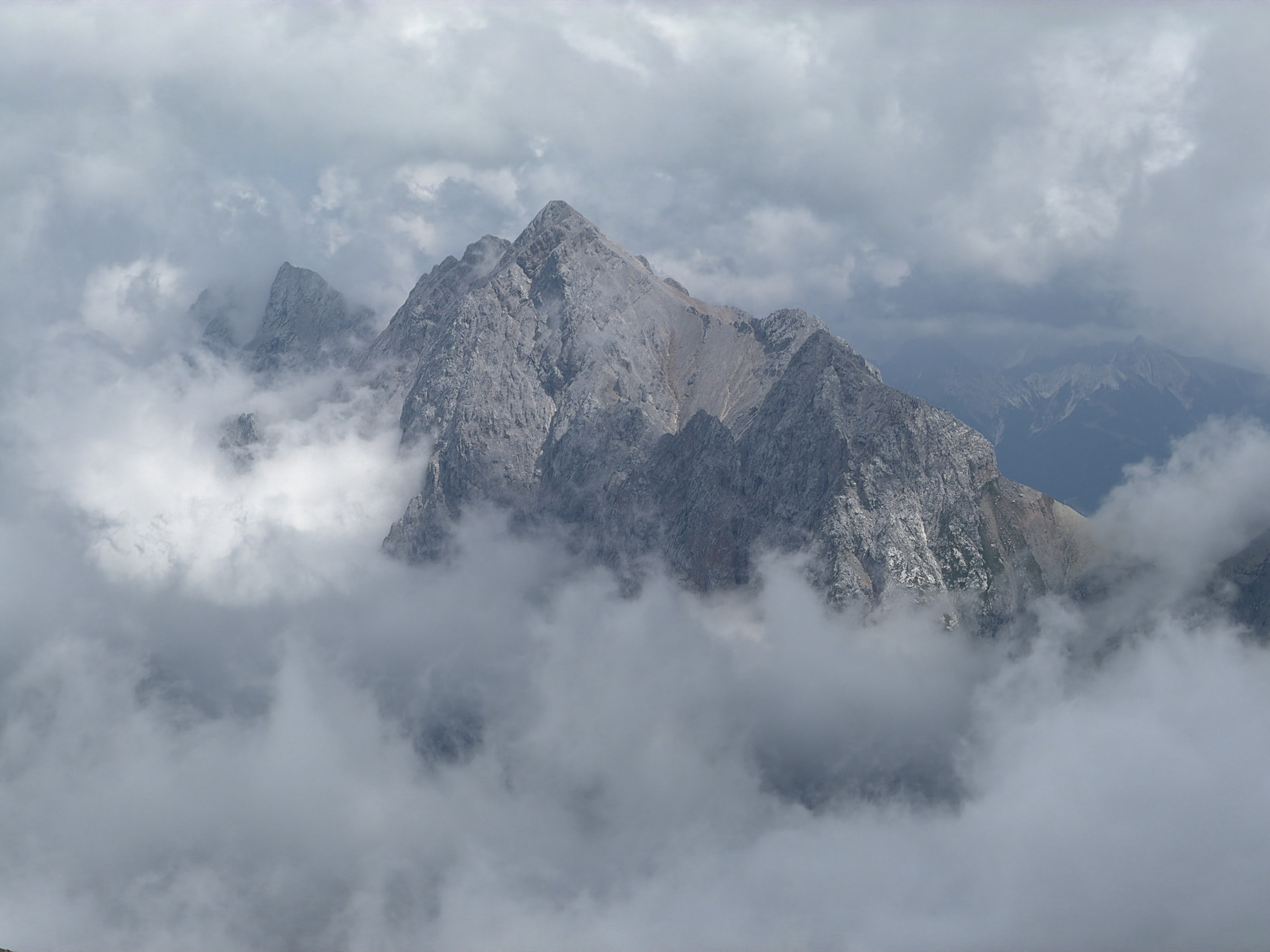 The image shows a dramatic mountain landscape, partially obscured by thick, swirling clouds. The prominent subject is a jagged, rocky mountain peak with a steep, imposing face. Its texture is rough and weathered, with visible striations and patches of scree. The summit is sharp and defined, piercing through the overcast sky. Surrounding this central peak, other mountain ridges and formations are visible, some peeking through the fog while others are completely enveloped. The clouds are dense and white, creating a soft, ethereal contrast with the harshness of the rock. The overall atmosphere is one of natural grandeur and a sense of being high above the earth. The location is the Alps, near Garmisch-Partenkirchen, Germany, likely depicting the Zugspitze or a surrounding peak. There are no people or man-made objects visible in the scene.
