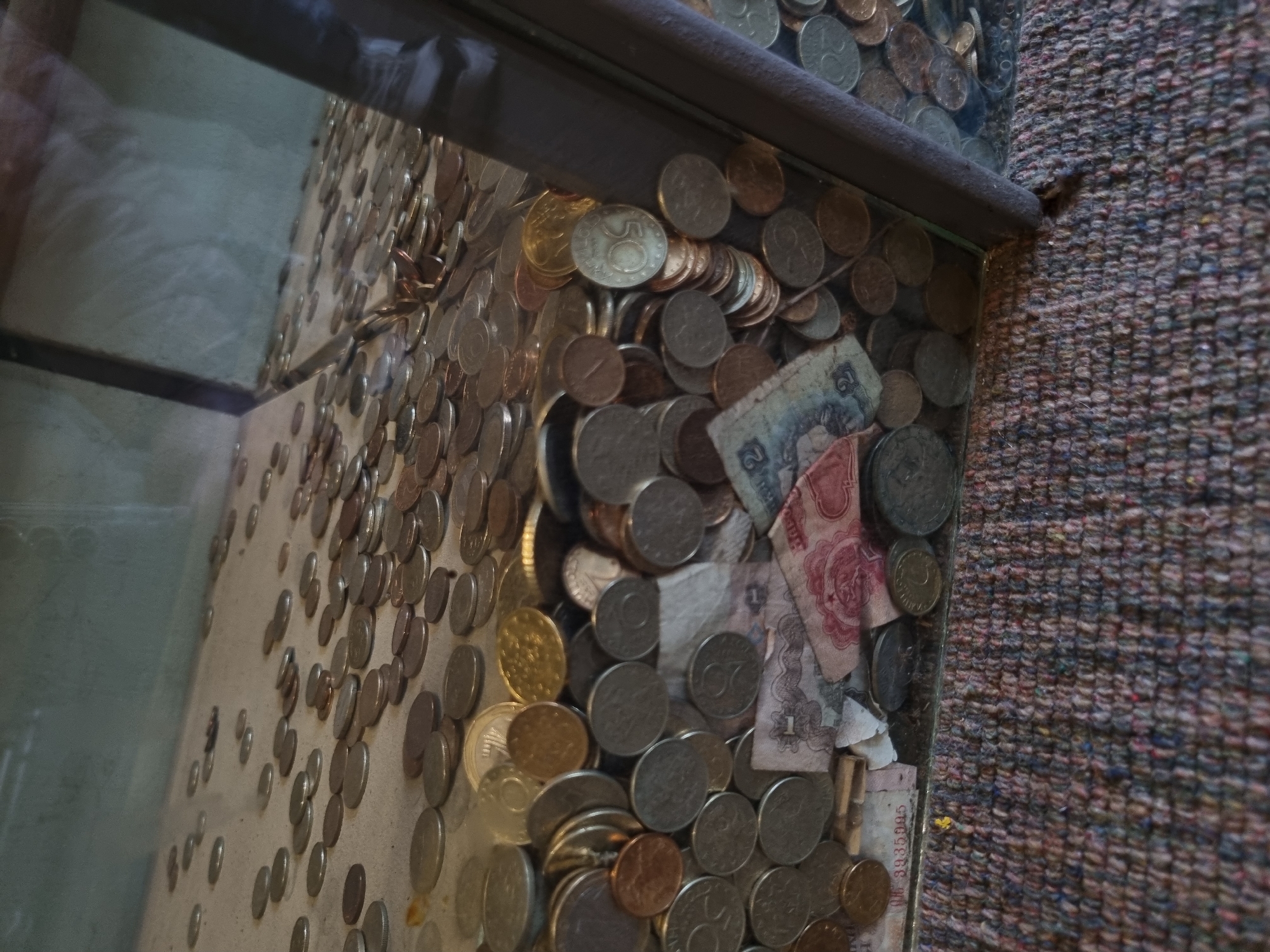 The image shows a glass donation box filled with coins and banknotes. The coins are various sizes and colors, with some appearing to be euro currency. The banknotes are crumpled and partially obscured by the coins. The box is positioned on a carpet. The location is Elena, Bulgaria.