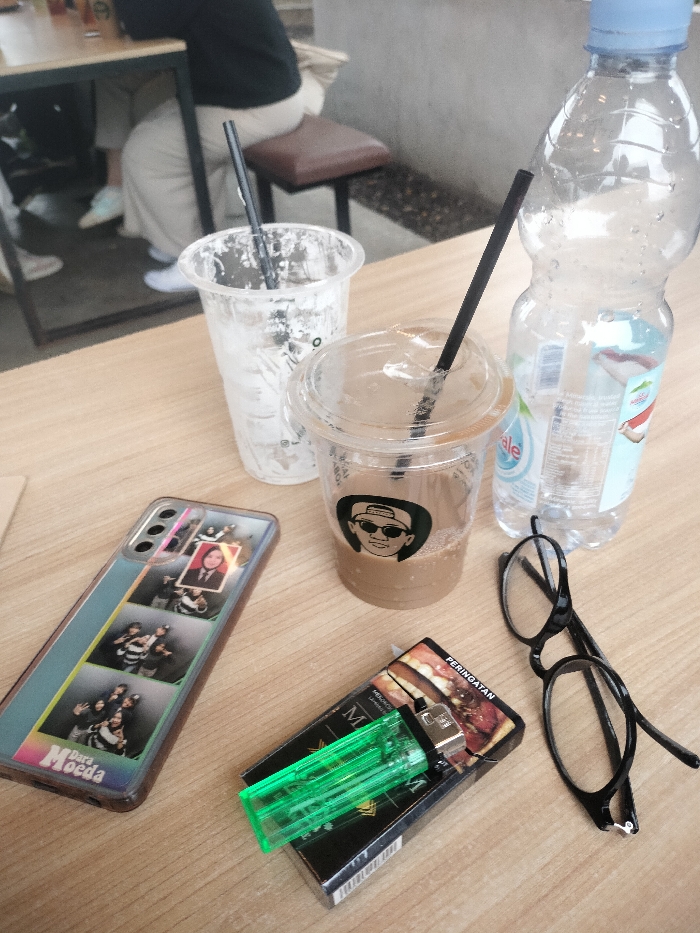 The image captures a wooden table holding various personal items and beverages in a casual, possibly outdoor or semi-outdoor, cafe setting in Dramaga, Indonesia.
In the foreground, a smartphone lies on the left, encased in clear plastic and featuring a collage of small photographs. Partial text "Para Moeda" is visible at the bottom of the phone case. To its right are two plastic cups with black straws. One cup is nearly empty with residual contents, while the other contains a partially consumed brown beverage, likely coffee, and displays a black and white logo of a person in sunglasses and a cap. A clear plastic bottle of "Le Minerale" water, mostly empty, stands beside the coffee cup.
Further to the right, a black pack of "MAGNUM" cigarettes is visible, bearing the Indonesian word "PERINGATAN" (Warning). A bright green plastic lighter rests on top of the cigarette pack. A pair of black-framed eyeglasses completes the collection of items on the right side of the table.
In the blurred background, two individuals are seated at another table. One person wears a black top and light-colored pants, with their legs and white shoes partially visible, suggesting a relaxed social gathering. The overall lighting is bright and even, indicating a daytime scene. The arrangement of items suggests a brief stop or break for drinks and possibly a cigarette.