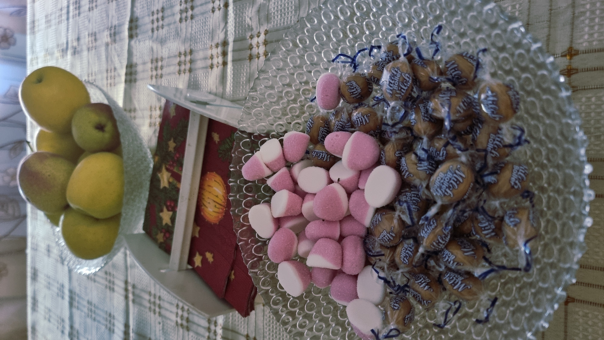 A close-up, overhead shot displays a clear glass bowl filled with assorted candies, primarily pink and white marshmallows and individually wrapped caramel candies, resting on a table covered with a checkered tablecloth. In the background, a glass bowl holds several pears. A festive red napkin with a gold star pattern is visible beneath the candy bowl. The scene is set on a table, suggesting an indoor environment, possibly a home or a catered event. There are no people present. The lighting is soft, casting subtle shadows. There is no information about Sant Joan Les Fonts, Spain.