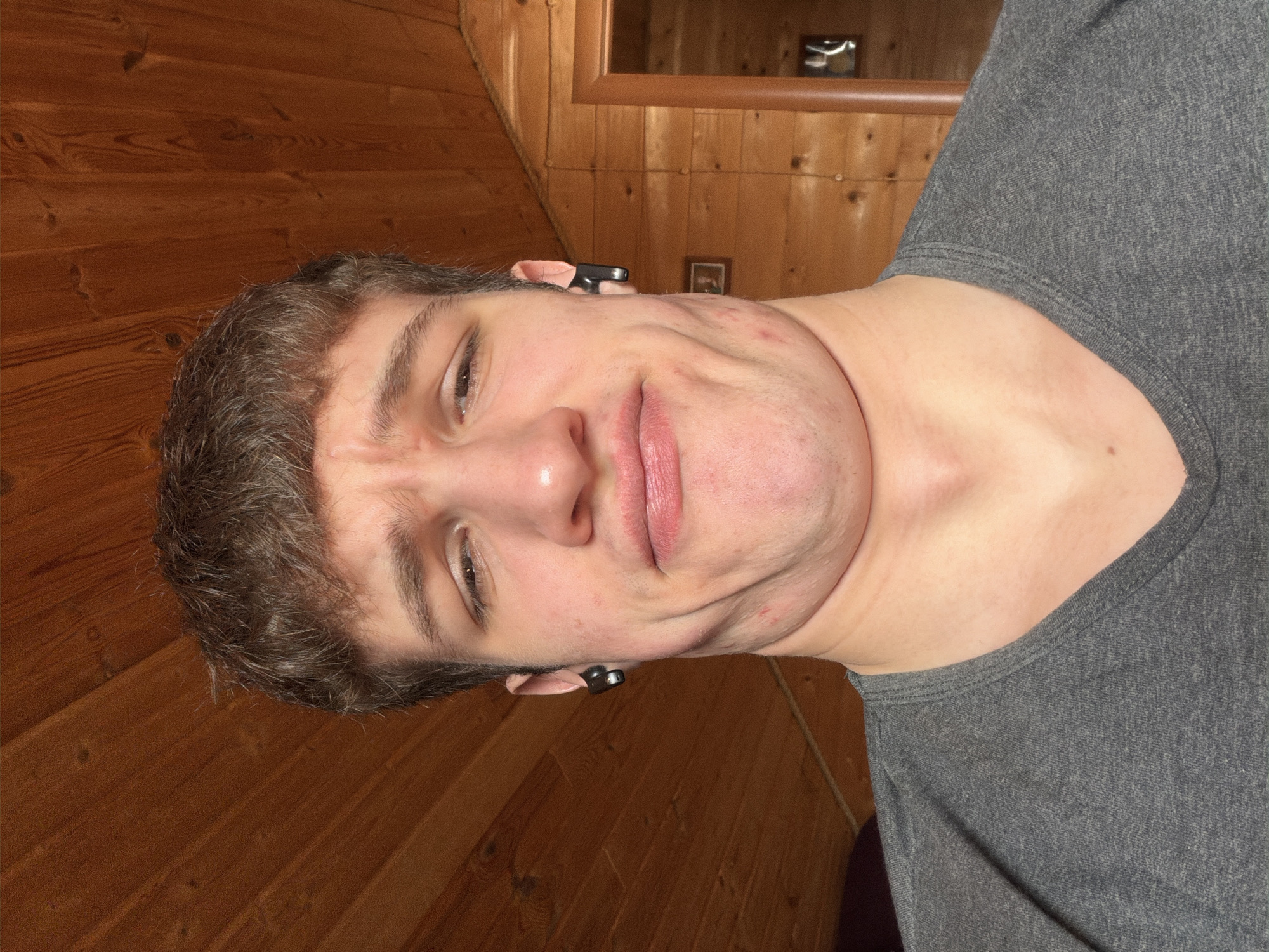 The image presents a close-up, rotated portrait of a young man, likely in his late teens or early twenties, with short, dark brown hair. He is looking directly at the camera with an exaggerated, playful expression, characterized by scrunched facial features and folds in his chin.
The man is wearing a simple grey crew-neck t-shirt. Small, black in-ear headphones are visible in both ears. His skin shows some visible blemishes on his forehead, cheeks, and chin.
The background consists of warm-toned wooden wall paneling, featuring prominent wood grain and knots, suggesting a rustic or homely indoor setting. A section of a wooden-framed mirror is visible in the upper right, reflecting a small, indistinct light source or object within the room. The lighting appears to be artificial and internal.
The overall mood is casual and informal, with the subject engaging the viewer with a humorous pose. The setting suggests a comfortable, private space, possibly in Прикамский, Russia. No visible text is present in the image.