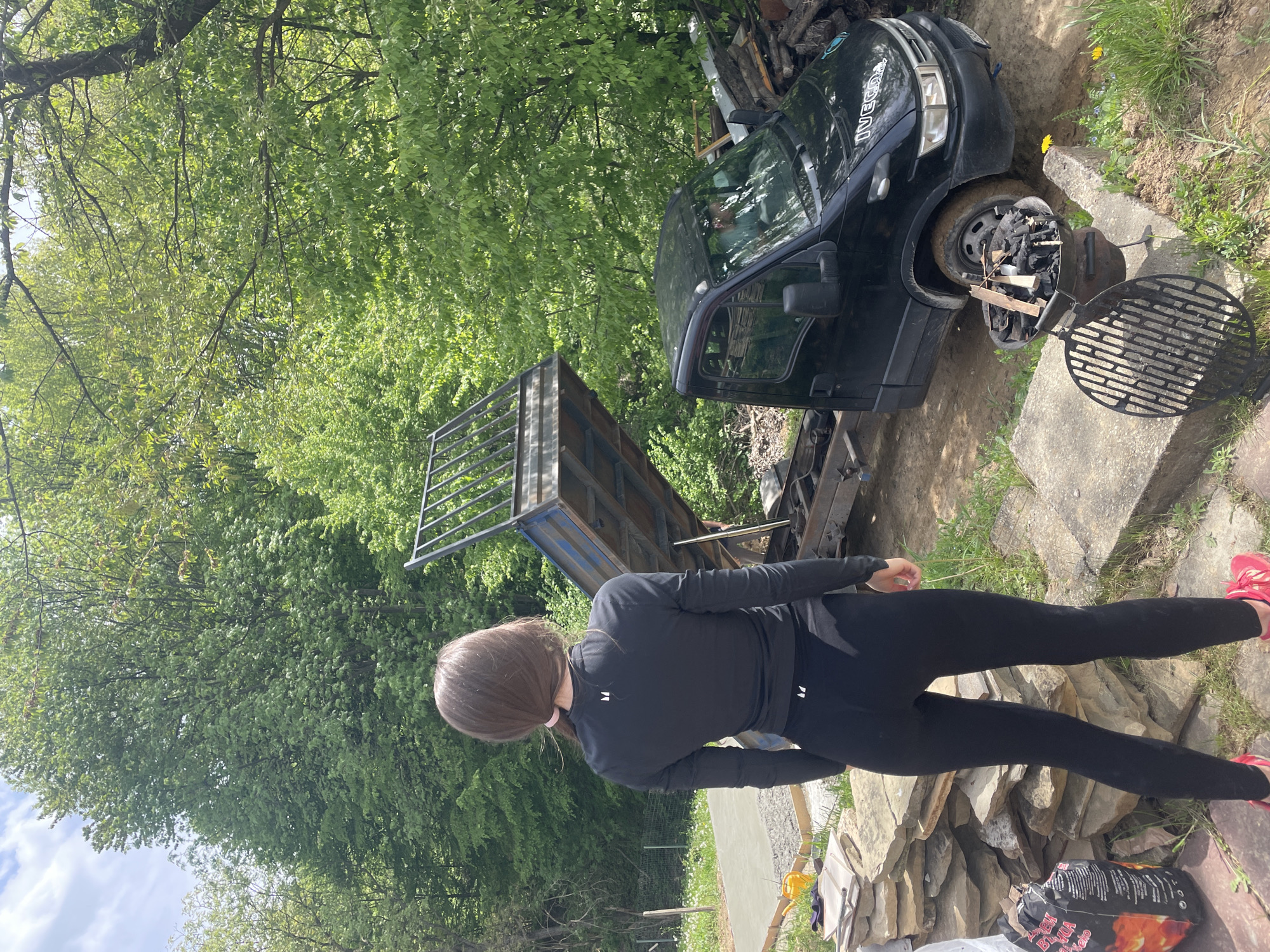 The image appears to be taken outdoors in Bogdanchovtsi, Bulgaria, possibly in a yard or garden area. A woman wearing a black long-sleeved shirt and black leggings stands with her back to the camera. To her left is a flatbed truck; behind it is a large green tree. To her right is a black car which is situated next to an outdoor grill. There are also stones visible in the foreground.