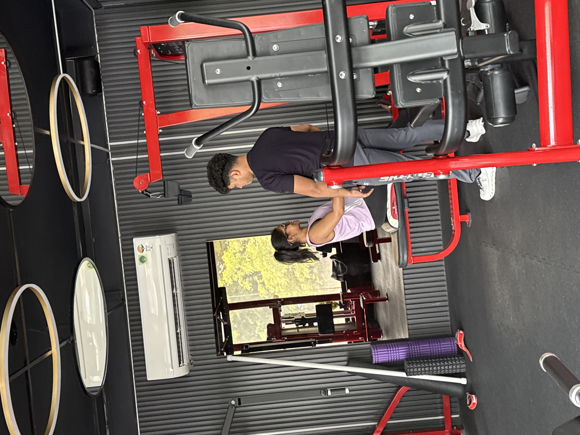 The scene depicts a gym setting, likely in Kolkata, India, featuring two individuals using a red, heavy-duty leg press machine. One person, wearing a black shirt and grey pants, is currently in a seated position on the machine, seemingly in the middle of a leg press exercise. The other individual, sporting a light purple shirt, appears to be assisting or observing the exercise.
In the background, a window overlooks foliage that suggests the gym is located in a somewhat green area. Other gym equipment, including rollers of purple and black colors, are scattered on the floor. An oval-shaped mirror and an air conditioning unit are visible on the wall. The walls are adorned with black and gray horizontal lines.