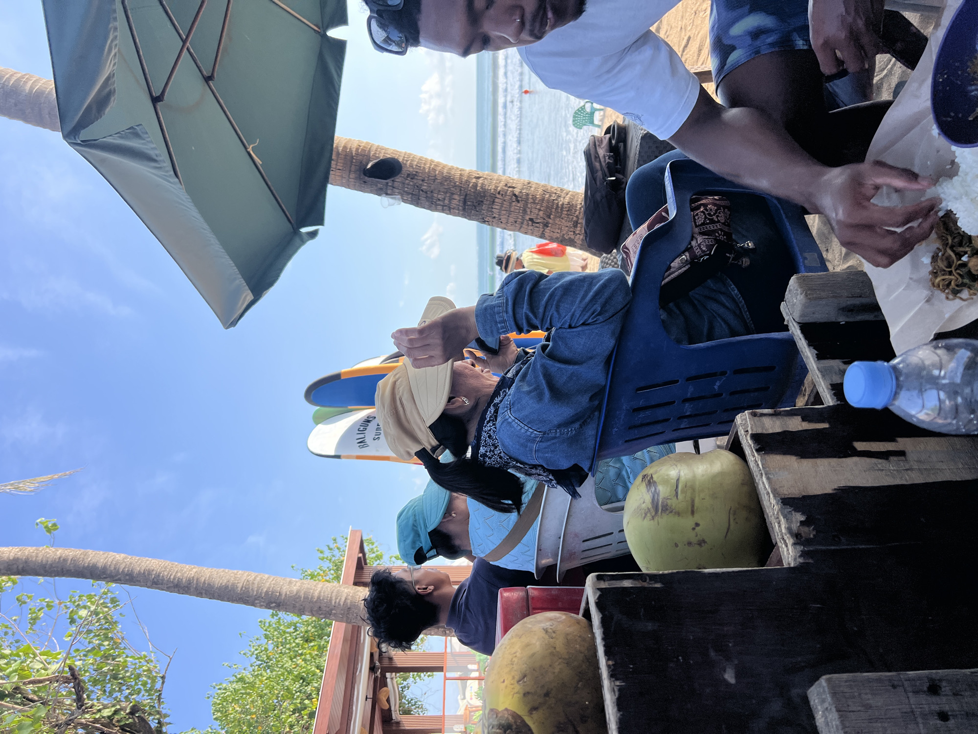 The image captures a scene at a beachside establishment in Kuta, Indonesia, likely a surf shop or cafe. Several people are seated under a large green umbrella, facing the ocean. In the foreground, on the left, are two coconuts resting in wooden crates. A man in a white t-shirt and patterned shorts is seated on the right, eating from a plate of food. To his left, a woman wearing a denim jacket and a tan sunhat is looking at her phone. Behind them, two other individuals are seated, one wearing a blue shirt and the other a darker top. Surfboards with "BALIGUNA SURF" branding are visible behind the seated individuals. The ocean is visible in the background with small waves breaking on the shore. The scene is bathed in sunlight, indicating a daytime setting.