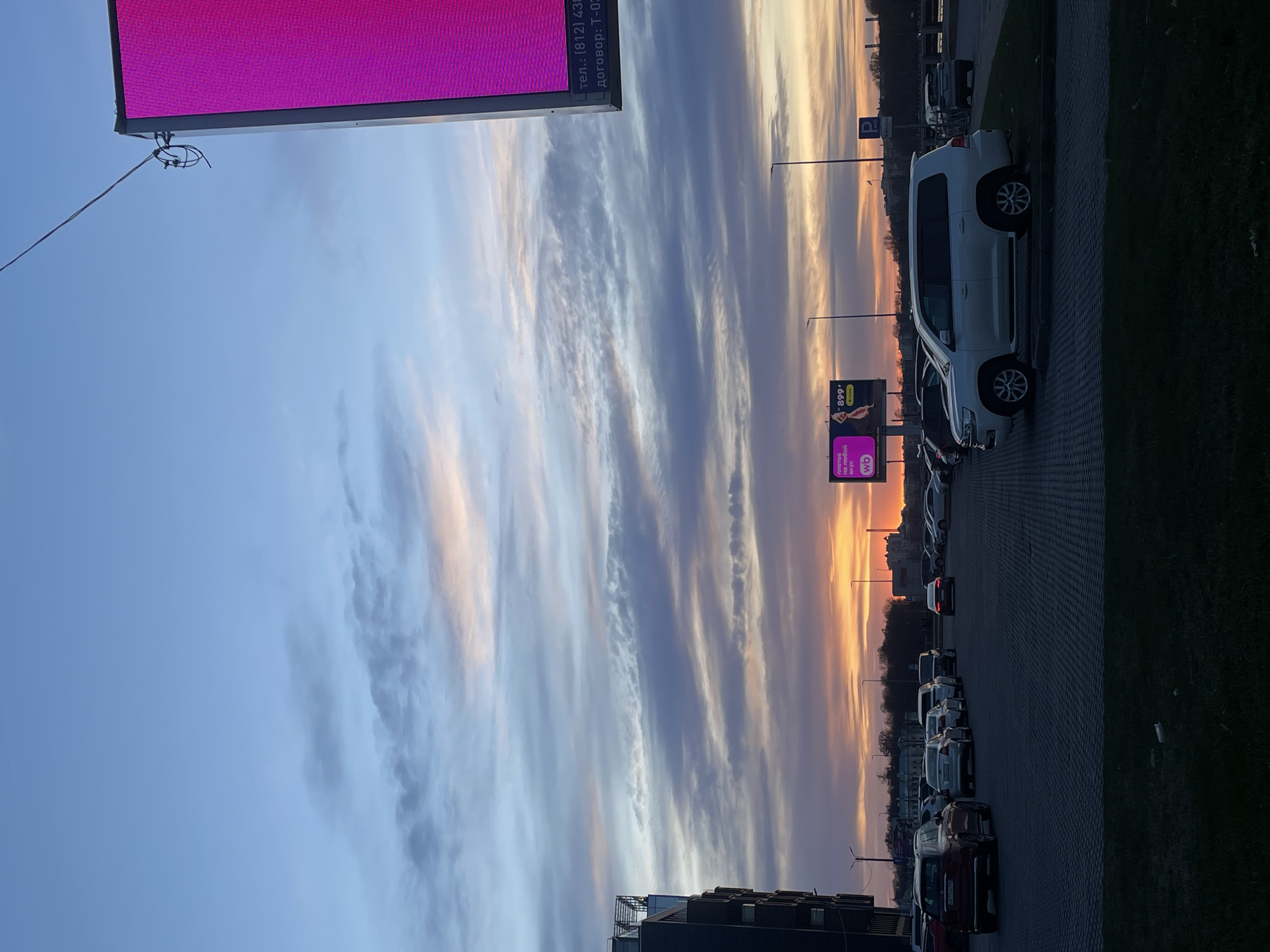 The image captures a street scene in Saint Petersburg, Russia, at dusk or dawn, with a distinctive sky gradient from blue to orange. A large billboard with a pink advertisement dominates the upper left. Alongside the road, numerous parked cars are visible, with a white SUV prominently positioned. A green strip of grass runs along the edge of the road, separating it from the parking area. A smaller advertising sign is seen further down the street. Buildings and structures are partially visible in the background. The image is oriented vertically.