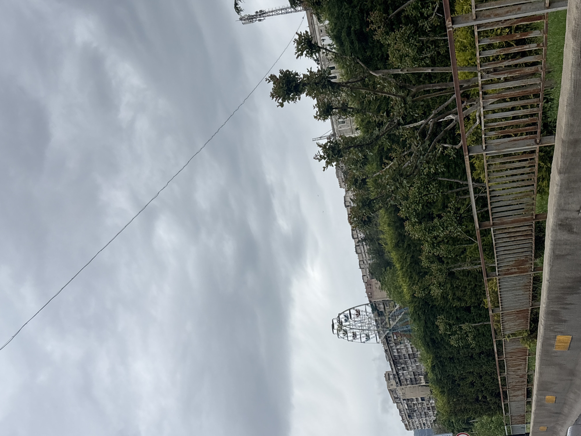 The image shows a cityscape in Tbilisi, Georgia on an overcast day. A rusty metal pedestrian bridge or walkway runs along the right side of the frame, partially obscured by greenery. Beyond the vegetation, tall buildings line the street. A Ferris wheel is visible in the distance on the left, indicating an amusement park or entertainment area. Above, the sky is filled with gray clouds. A power line stretches across the upper left portion of the image.
