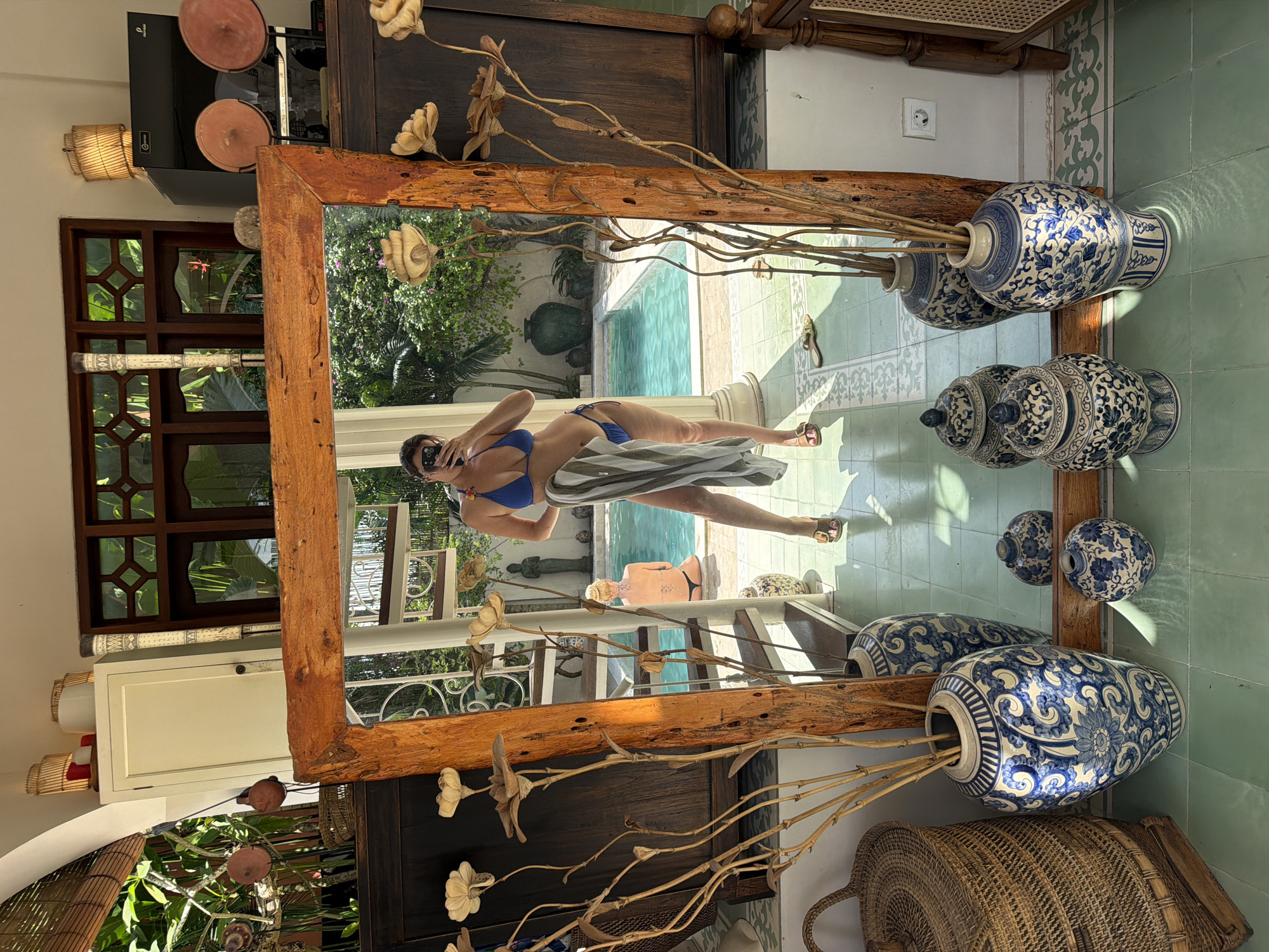 A large, rustic wooden-framed mirror is the central feature of this image, positioned at an angle on a light green and white patterned tile floor. In the mirror's reflection, a woman is seen taking a selfie with her phone, wearing a blue bikini and sunglasses, with a striped towel draped over her lower body. The reflection reveals an outdoor scene with a bright blue swimming pool, lush green foliage, and what appears to be a stone statue. Another person, bald and light-skinned, is partially visible lying near the pool. A pair of sandals are also reflected on the poolside.
The interior space surrounding the mirror features several decorative elements. To the right of the mirror, two large, blue and white patterned ceramic vases with dried branches are displayed, along with two smaller, matching lidded jars. On the left side, dried floral arrangements, possibly made of carved wood, adorn the area. Visible beyond the mirror's frame are dark wooden furniture, a woven wicker basket on the floor, and a wicker light fixture mounted on a wall. Above a wooden window frame with decorative lattice work, a dark flat-screen monitor or TV with "Proview" faintly visible on its screen is mounted, with two terracotta-colored decorative plates on a shelf below it. A white electrical outlet with a yin-yang symbol is also seen on the wall, as is a white cabinet and a long, decorated cylindrical object.
The scene is brightly lit by natural sunlight, indicating daytime. The lush greenery, swimming pool, and architectural style suggest a tropical or resort-like setting, consistent with its North Kuta, Indonesia location.