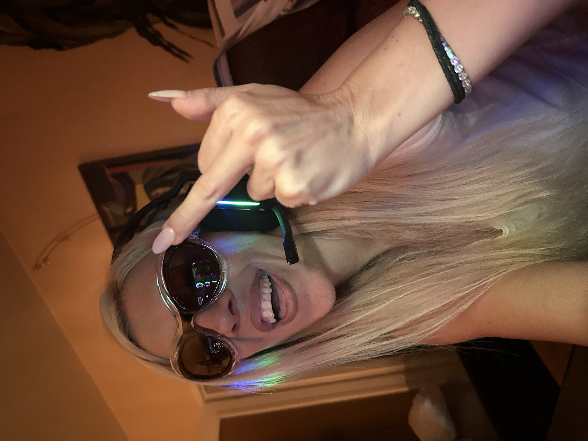 A young adult female with blonde hair is pictured from the chest up, indoors. She is wearing large, novelty sunglasses with clear frames that refract light in rainbow hues. She has on a headset with a microphone extending from the right side. Her mouth is open in a wide smile, revealing her teeth. Her right hand is raised, and she is extending her middle finger towards the camera. She has long, light-colored fingernails. On her left wrist, she wears a black and crystal bracelet. The background is dimly lit with warm-toned lighting, suggesting an indoor evening setting. There are hints of furniture and possibly a painting on the wall behind her. The specific location in Pazardzhik, Bulgaria, cannot be determined from the image.