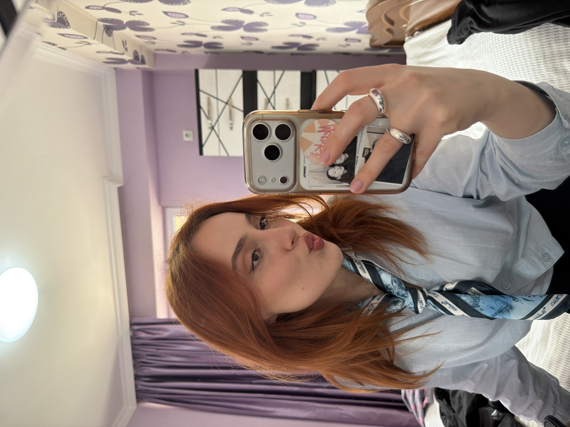 A young woman with red hair takes a selfie in an interior space. She is wearing a light blue button-down shirt and a blue and white patterned scarf. She holds a phone with a gold-colored case, stickers, and images attached to it. She has silver rings on her fingers. The background includes light purple walls, a white headboard with a purple floral pattern, and a modern black and white wardrobe. The specific location in Tbilisi, Georgia, is not discernible from the image.