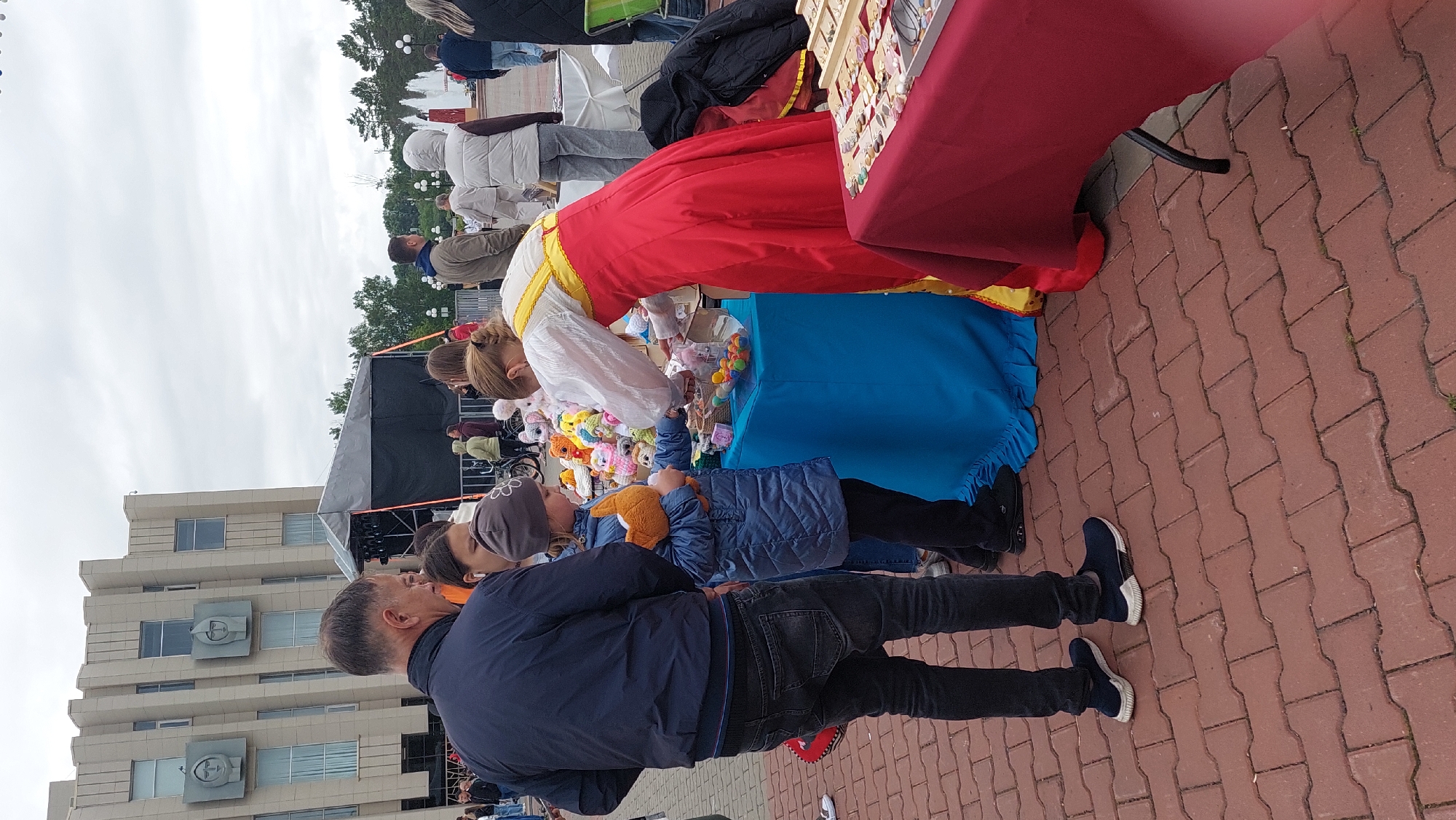 In Komsomolsk-on-Amur, Russia, a street scene unfolds near a Brutalist-style building with decorative reliefs. A group of people are gathered around two tables displaying various items.
In the foreground, a man in a dark blue jacket and jeans stands with his back to the camera, looking towards a stall. Beside him, a young girl in a blue jacket and a grey beanie holds a stuffed orange fox. She is interacting with a woman dressed in a traditional red and yellow outfit, who is tending to a table covered with a blue cloth. This table has a clear container filled with colorful candies and several small, plush toys.
To the right of this stall, another table draped with a red tablecloth displays an assortment of jewelry, including necklaces and earrings. In the background, other individuals are visible, some walking and others observing the stalls. A stage with black draping is set up further back, suggesting an outdoor event or festival. The ground is paved with reddish-brown brick pavers. The sky is overcast.