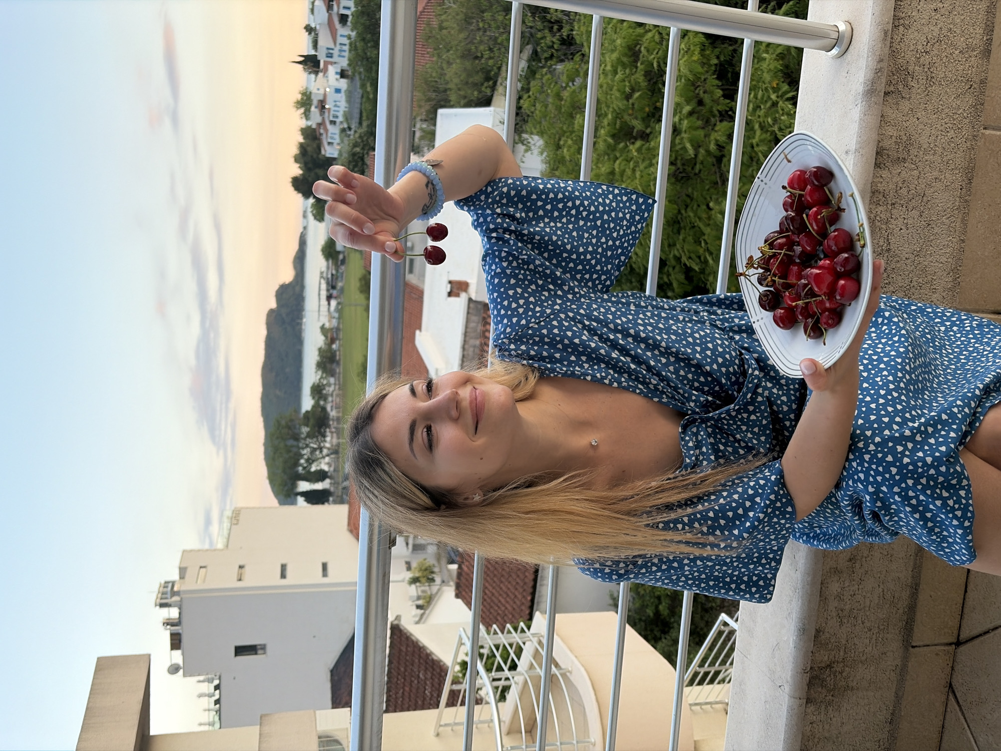 On a balcony in Budva, Montenegro, a fair-skinned woman with long blonde hair poses with a bowl of red cherries in her right hand and two cherries dangling from her left hand. She is wearing a short-sleeved blue dress with white heart patterns. The balcony railing is white. In the background, there are trees, white buildings with red tile roofs, a body of water, and a sky with a mix of blue, white, and orange hues.