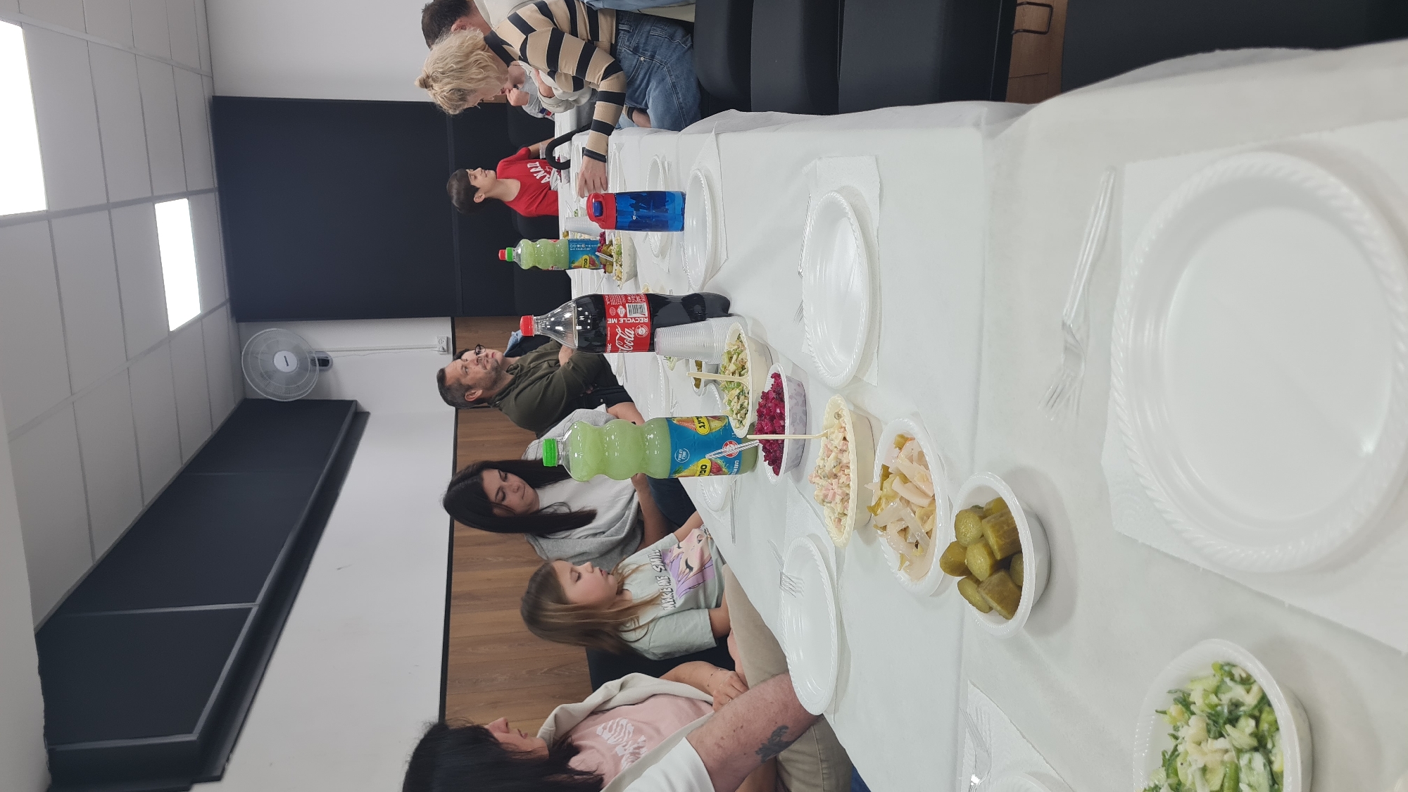 A photograph taken indoors shows a group of people seated around a long table covered with a white tablecloth. The table is set with white disposable plates, forks, and small bowls containing various salads. Several bottles of soda and water are also placed on the table.
Individuals of varying ages are seated at the table, engaging in conversation. The setting suggests a casual gathering, possibly a meal or celebration. The indoor location appears to be a simple meeting room, possibly in a community center. A black object is placed under a fan on the wall.
While the photograph gives clues about the location, the city of Nahariyya, Israel, cannot be confirmed from the information provided.