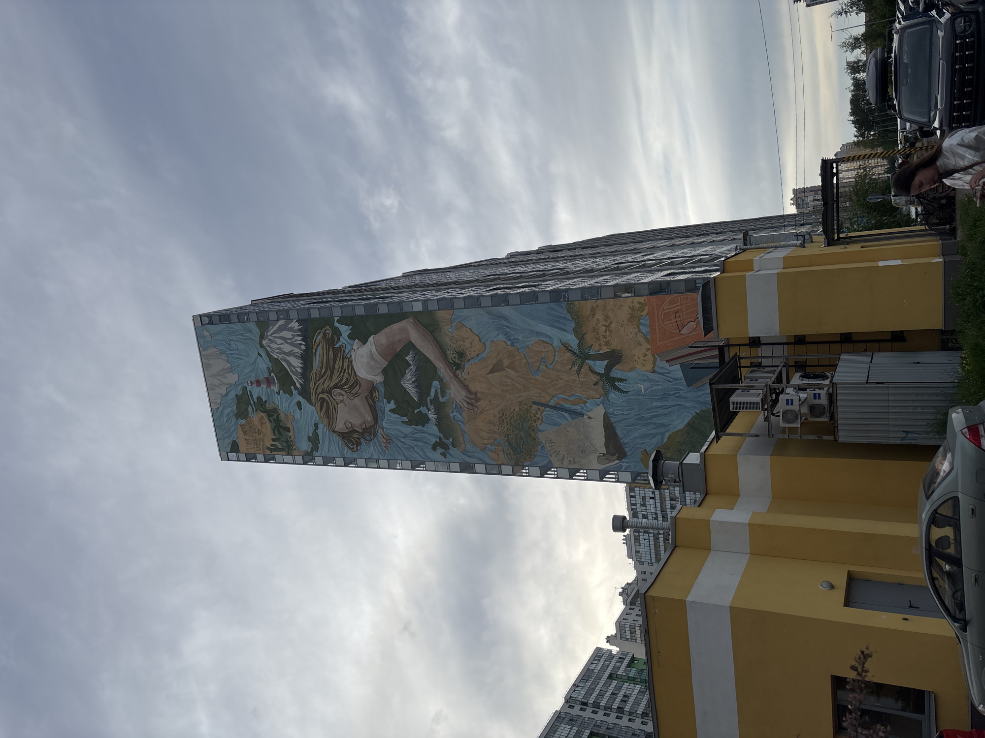 A tall, narrow building stands prominently, featuring a large mural painted across its side. The mural depicts a stylized map, likely of the world or a major continent, rendered in shades of blue for water and earthy tones for landmasses. A central figure, a woman with long, flowing blonde or light-colored hair and a serene expression, is integrated into the map, her arm extending over what appears to be a body of water. Elements like an old sailing ship are visible on the map.
In front of this mural-adorned building, a smaller, yellow building with two horizontal white stripes is visible. Air conditioning units are mounted on its roof. Various vehicles are present; a grey car is partially seen in the foreground, and a dark SUV or truck is parked further back. Other residential buildings are visible in the distance, suggesting an urban or suburban setting. Overhead wires stretch across the scene.
The sky is overcast or partly cloudy, indicating daytime with diffused light. No specific activity is occurring beyond the static presence of buildings and parked cars. A person with dark hair, wearing a light-colored top, is faintly visible in the upper right corner of the overall scene, near the parked cars, with their back mostly to the viewer. There is no visible text.