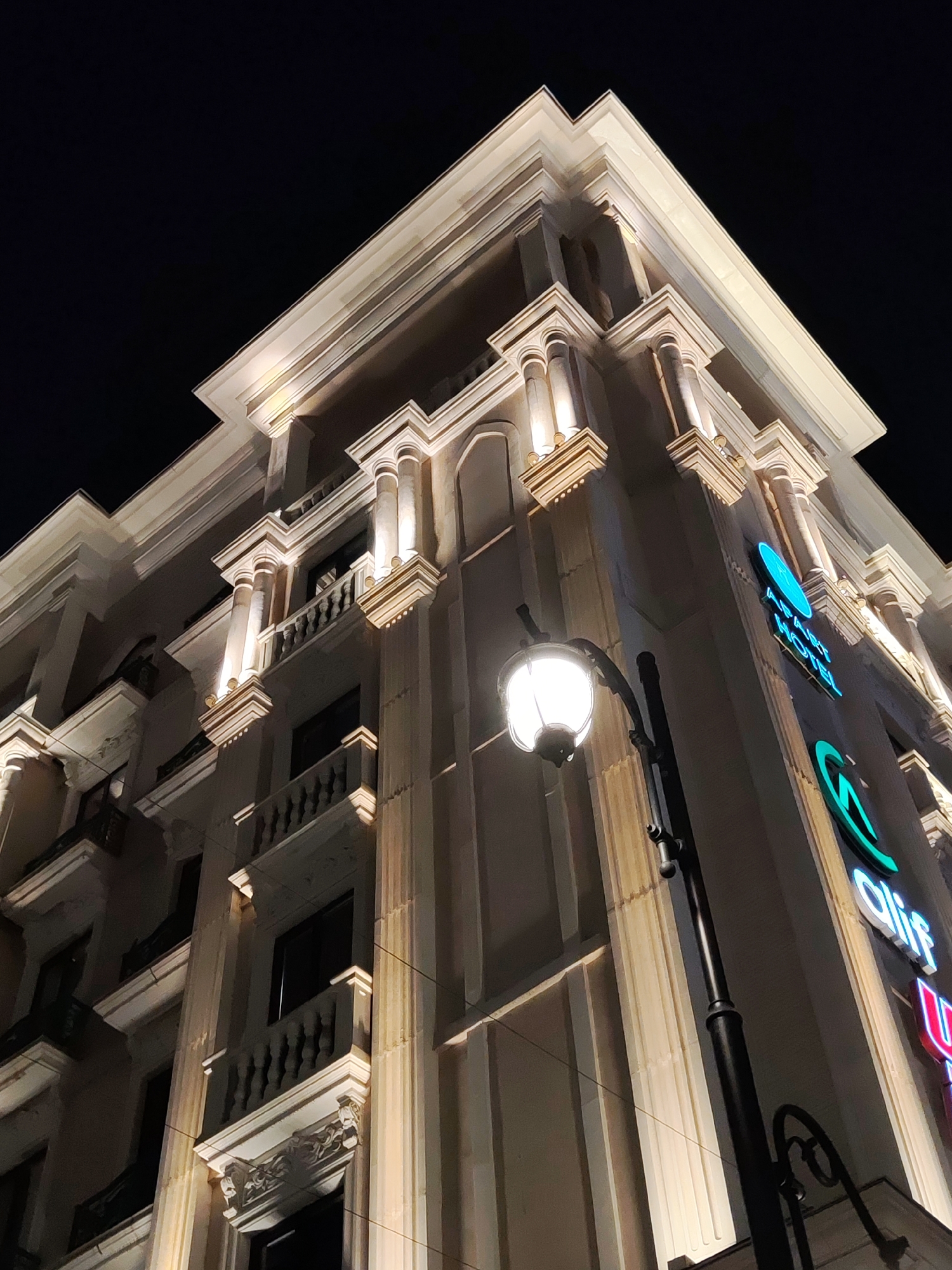 The image captures the exterior of a modern hotel in Tashkent, Uzbekistan, at night. The building features a classical architectural style with illuminated columns, balconies, and decorative elements. A street lamp with a spherical light fixture stands in the foreground, casting a warm glow. Neon signs, including "ALIF HOTEL," are visible on the building's facade. The sky is dark, indicating the time of day. The overall scene is well-lit, highlighting the building's design and its nighttime ambiance.