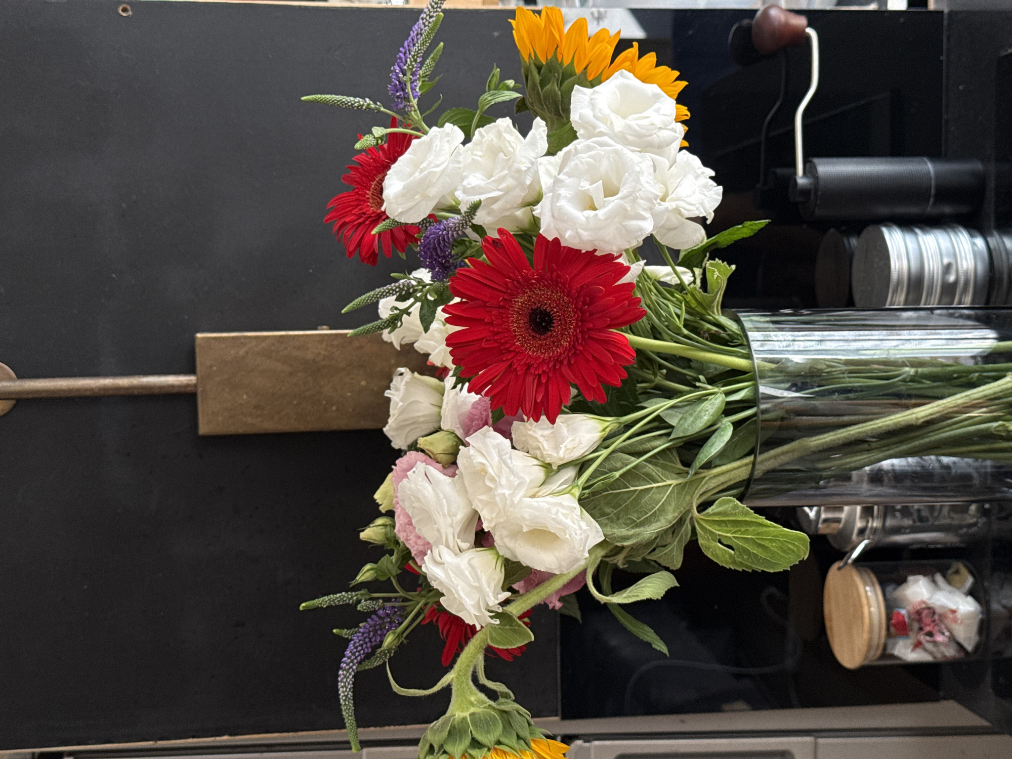 A close-up, high-angle shot captures a vibrant bouquet of flowers in a glass vase, positioned against a dark, textured background. The arrangement features prominent red gerbera daisies, interspersed with delicate white lisianthus, and accented by sprigs of purple veronica and hints of pink. A partially visible sunflower adds a touch of yellow to the left, while a bright orange sunflower peeks from the right. The flowers are in various stages of bloom, with some buds still closed. The vase, filled with water and the flower stems, rests on a dark surface, likely a countertop. To the left of the vase, a clear glass jar with a wooden lid contains small, wrapped items. Next to it is a metallic, cylindrical object, possibly a shaker. To the right of the vase, a sleek, black manual coffee grinder with a silver crank and a wooden handle stands upright. The overall setting suggests an indoor environment, potentially a kitchen or dining area in Tel-Aviv, Israel.