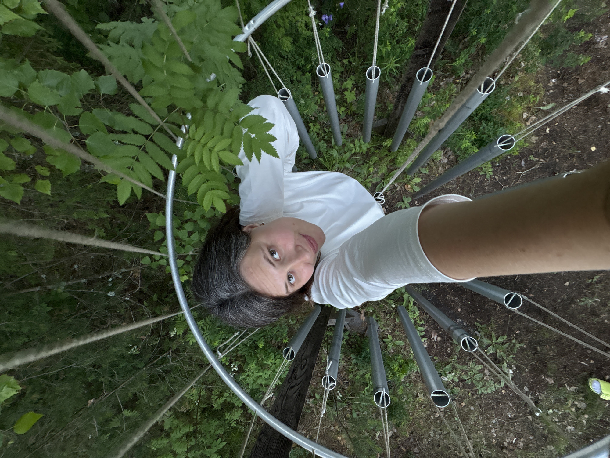 A woman is seen from a high angle, looking up and taking a selfie. She has dark hair with some gray streaks and is wearing a white t-shirt. She is standing on a spiral staircase made of metal pipes and ropes, which is surrounded by dense green foliage. The staircase appears to be in a wooded area, possibly a park or natural reserve. The ground below is covered with fallen leaves and some small plants. No specific location in the city of Kostovaty, Russia can be identified from the image.