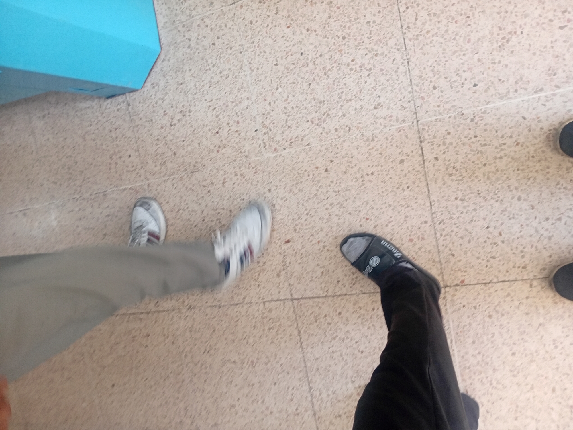 The image shows a low-angle view of two individuals' legs and feet standing on a terrazzo-patterned floor, likely indoors, with a portion of a light blue, angular object on the right side of the frame. The person on the left is wearing black pants and a black sandal with a grey sock peeking out. The sandal has "Zema" written on it. The person on the right is wearing light gray pants and white athletic shoes with red and blue stripes. The shoes are slightly blurred, suggesting movement. The floor is made of light-colored tiles with small, dark speckles. The blue object appears to be a counter or kiosk, possibly in a public space such as a store or office. The scene is well-lit, with the light source likely coming from above and slightly to the right. The image does not provide specific clues to pinpoint the exact location within Onikişubat, Turkey, but the flooring and the type of blue structure are common in commercial or public buildings.