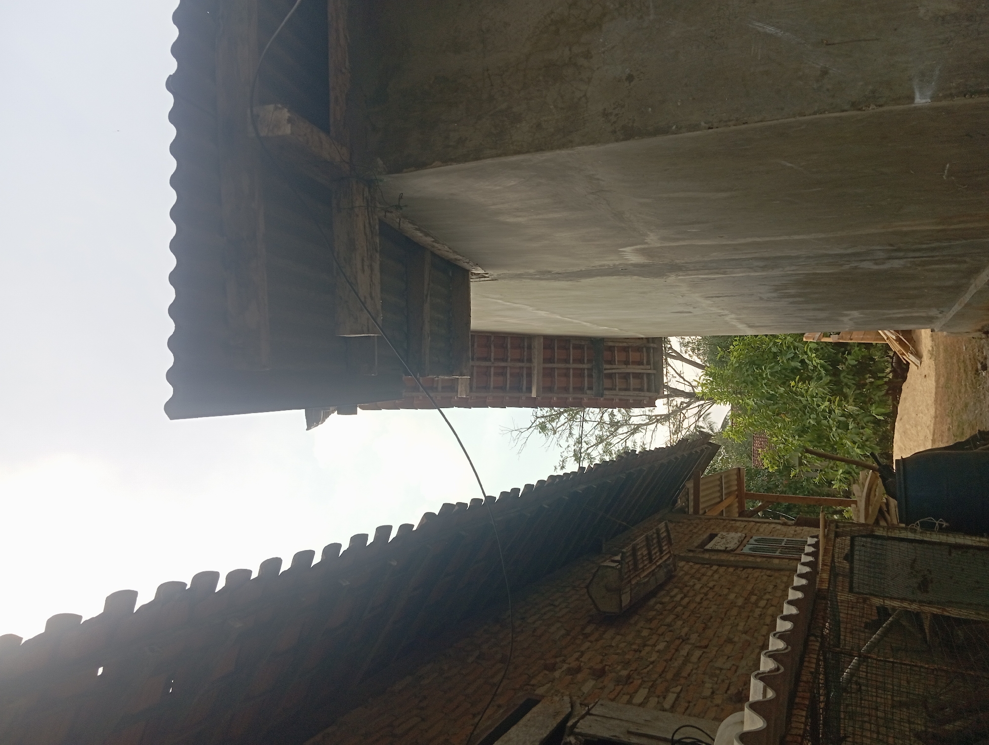The image shows a close-up view of two adjacent structures, possibly houses, in what appears to be a rural or semi-rural setting in Dente Teladas, Indonesia. The building on the left is constructed of red brick and has a roof made of terracotta tiles. The building on the right has a wall made of concrete, with a corrugated metal roof supported by wooden beams. The sky is overcast. Green foliage and trees are visible in the background, suggesting an outdoor environment. There are no people or animals visible in the image.