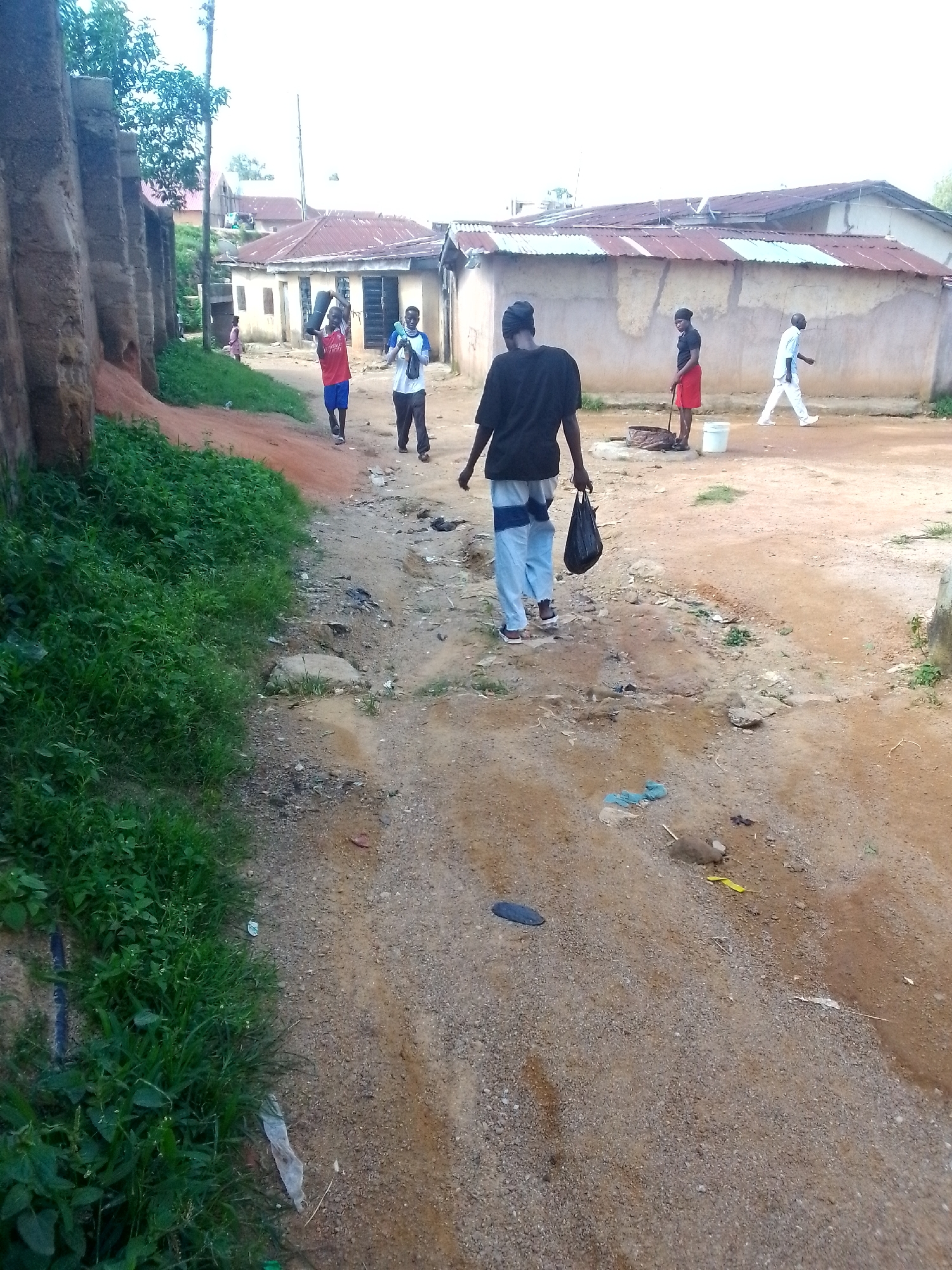 The image depicts a dusty, unpaved road in a village or town, with low-lying buildings with corrugated metal roofs on either side. Several people are present, walking or standing. On the left, a person in a red shirt and blue shorts is carrying a dark, cylindrical object over their shoulder. To their right, another person in a blue and white shirt and dark pants walks past. Further down the road, a person in a black t-shirt and light blue and white striped pants walks away from the camera, carrying a black plastic bag. To the right of the road, a person in a black shirt and red skirt stands near a well-like structure and a white bucket. Another person in a light blue shirt and white pants walks away from the well area. The ground is covered in dirt, gravel, and some scattered debris, including plastic bags and what appear to be discarded items. Green vegetation is visible along the left side of the road. The sky is overcast.