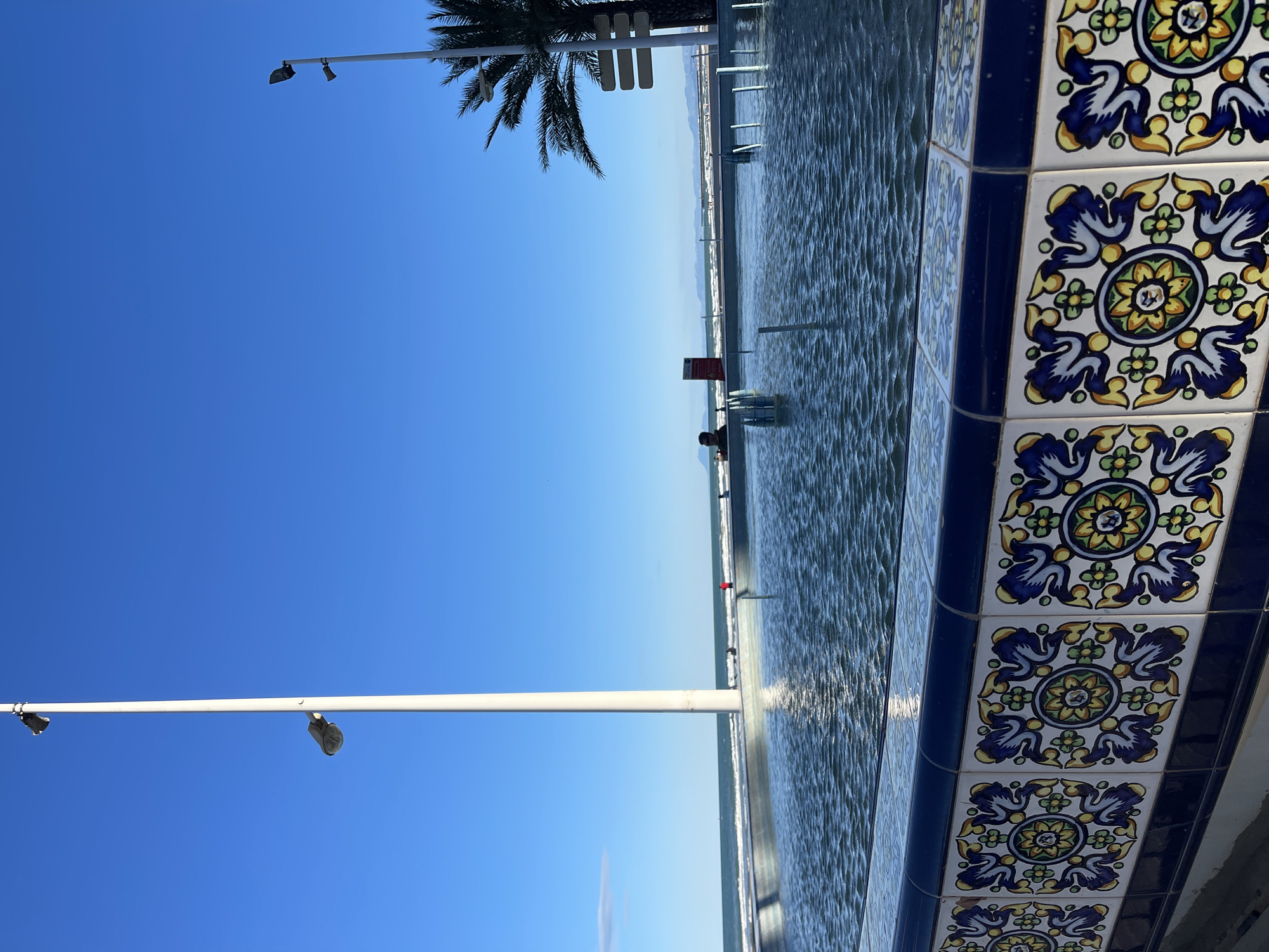 A vertically oriented outdoor shot taken in Cullera, Spain, depicts the beachside environment. The composition features decorative tiling in the lower right corner, with a white, light-colored wall beneath. The water’s edge extends across the frame, reflecting light with a rippling effect. A lifeguard station and distant beachgoers are visible along the waterline. To the left, a clear blue sky dominates, punctuated by a few light standards and the fronds of a palm tree in the top center.