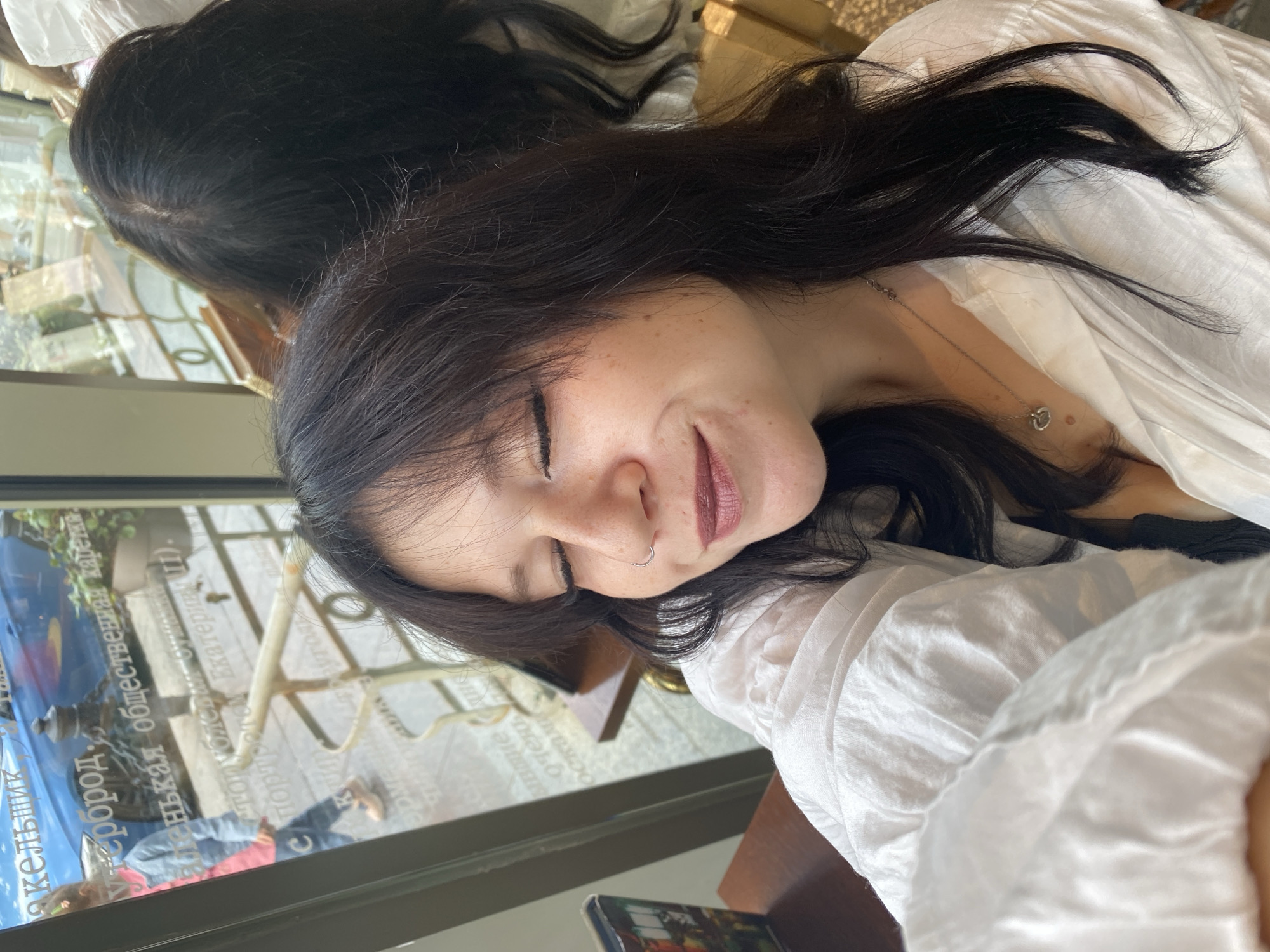 In a close-up selfie taken indoors, a young woman with dark hair and closed eyes smiles gently. She is wearing a white button-down shirt, a silver necklace, and a nose ring. The background, visible through a window, shows reflections and glimpses of the city of Saint Petersburg, Russia. The window displays text in Cyrillic, suggesting the location is a public establishment. A child with a pink and blue jacket and light brown shoes is walking outside, and a blue car is parked nearby. The scene also includes glimpses of indoor furniture, a potted plant, and a railing.