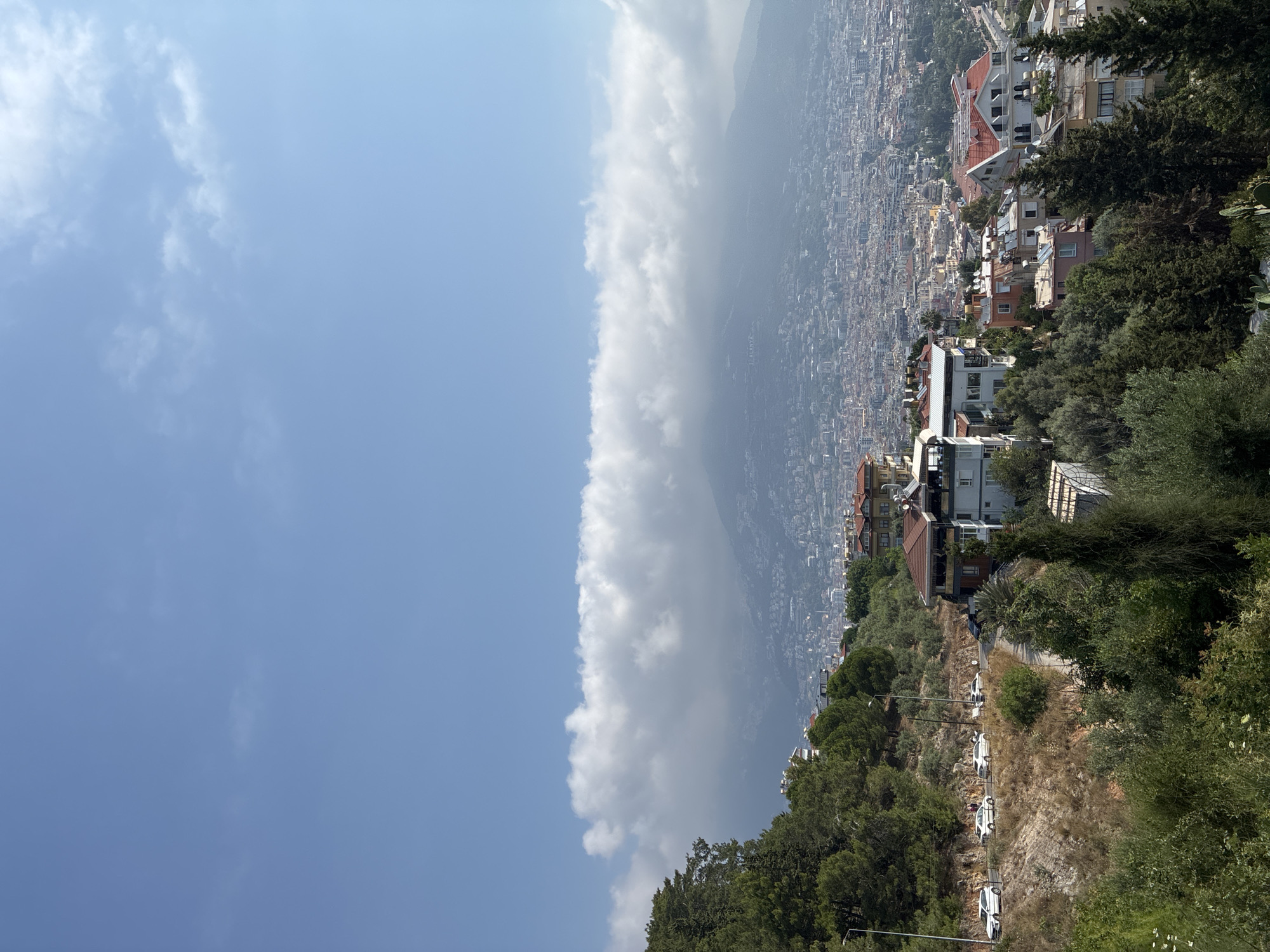 The image shows a panoramic view of Alanya, Turkey, from an elevated position, likely a hillside overlooking the city. In the foreground, there is lush green vegetation, including trees and shrubs, and a winding road with several cars. Some houses are visible on the slopes.
The middle ground is dominated by a dense urban landscape of Alanya, with numerous buildings of varying heights and architectural styles, many featuring red-tiled roofs. The city stretches towards the background, where a mountain range is visible. A distinctive band of white clouds is positioned horizontally across the sky, partially obscuring the upper reaches of the mountains. The sky itself is a clear, pale blue. The overall impression is one of a sunny day in a Mediterranean coastal city.