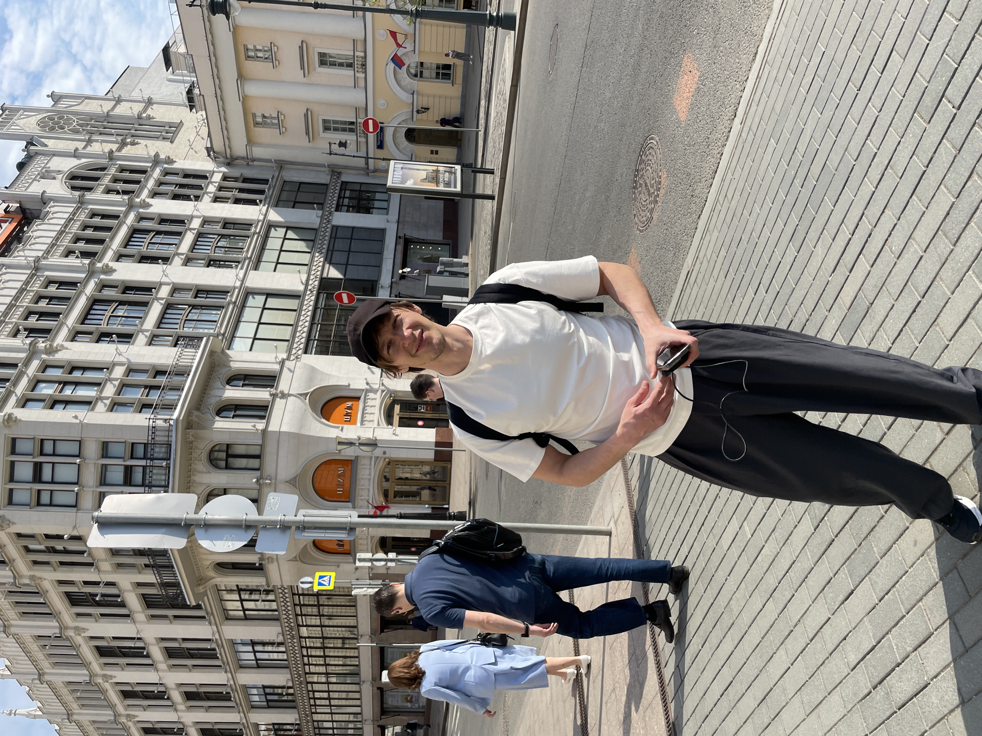 The image captures a street scene in Moscow, Russia, where a young man in a white t-shirt and black pants stands on a brick-paved sidewalk, holding a smartphone. He wears a black cap and a backpack. Behind him, pedestrians walk along the sidewalk. The background showcases Moscow's architecture, featuring a multi-story building with arched windows and signage written in Cyrillic. The buildings across the street exhibit a mix of architectural styles, with signage indicating commercial establishments. Street signs and traffic signals are also present, along with a Russian flag visible on one of the buildings.