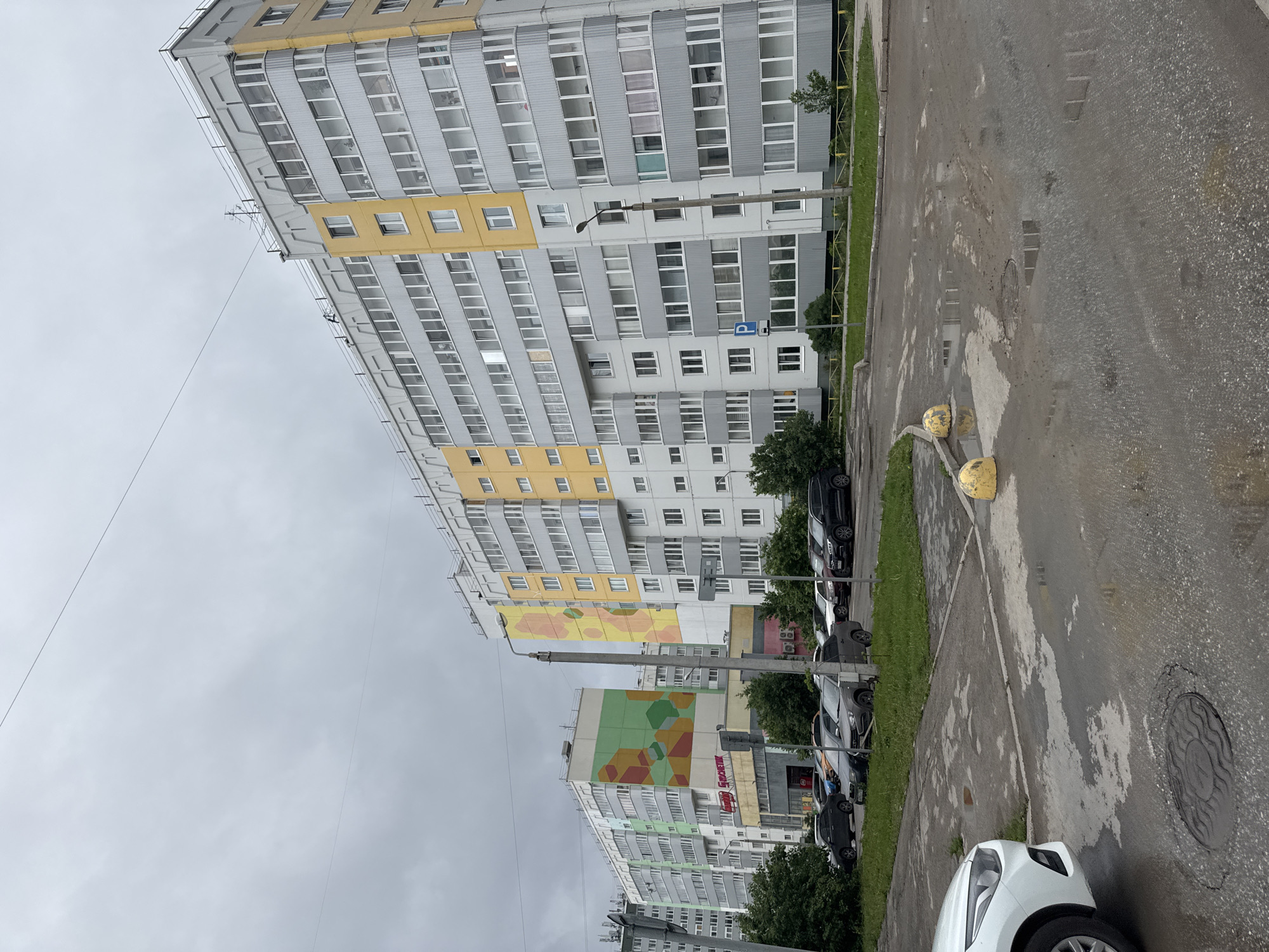 This image, presented rotated ninety degrees clockwise, captures a residential street scene in Kondratovo, Russia, under an overcast sky. The ground is wet with visible puddles on the asphalt road and sidewalk, suggesting recent rain.
The primary subjects are several multi-story apartment buildings. The most prominent building in the foreground is tall, featuring a grey facade interspersed with yellow horizontal bands and numerous windows. Another building further down the street exhibits a large, colorful abstract mural in shades of green, orange, and yellow. A third building is visible in the background, also appearing to have some colorful elements.
Along the roadside, numerous cars are parked, including a white car in the immediate foreground and several dark-colored vehicles further away. There are green grass verges separating the road from the buildings, and short fences are visible. Streetlights stand between the buildings and the parked cars. On the ground, two prominent yellow spherical bollards are visible, and a manhole cover with a decorative pattern is present in the asphalt. Overhead, thin electrical wires stretch across the cloudy sky.
Notable details include the dull, gray light indicating an overcast day. A blue square sign with a white "P" (parking sign) is affixed to a pole near one of the buildings. On the building with the green mural, a red sign with white text is partially visible, appearing to spell "МЕБЕЛЬ" (Mebel, meaning furniture). The architecture and overall setting are characteristic of modern residential developments in Russia. No people or specific events are depicted, providing a static view of the urban environment.