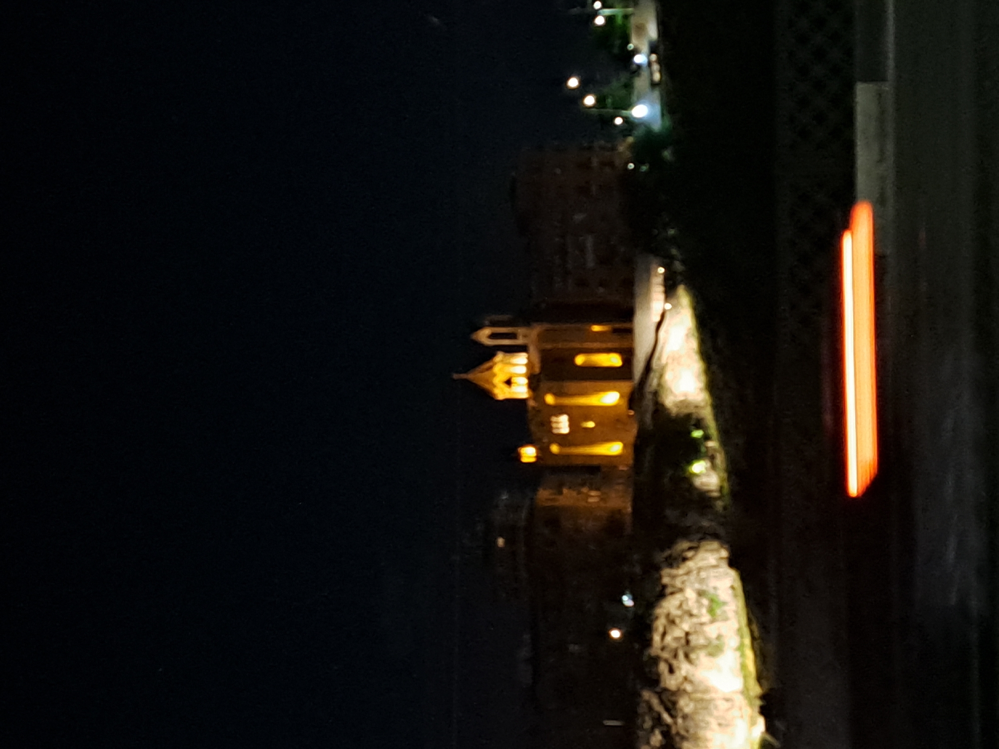 At night, a view from what appears to be a tall building overlooking Yerevan, Armenia, displays a building illuminated in yellow with a steeple, which might be a church. The scene includes a rocky terrain area near the building. To the right, there is an area of green, potentially trees or a garden. A vertical light source is seen, possibly part of the building or other lighting elements in the city. The rest of the background is dark.