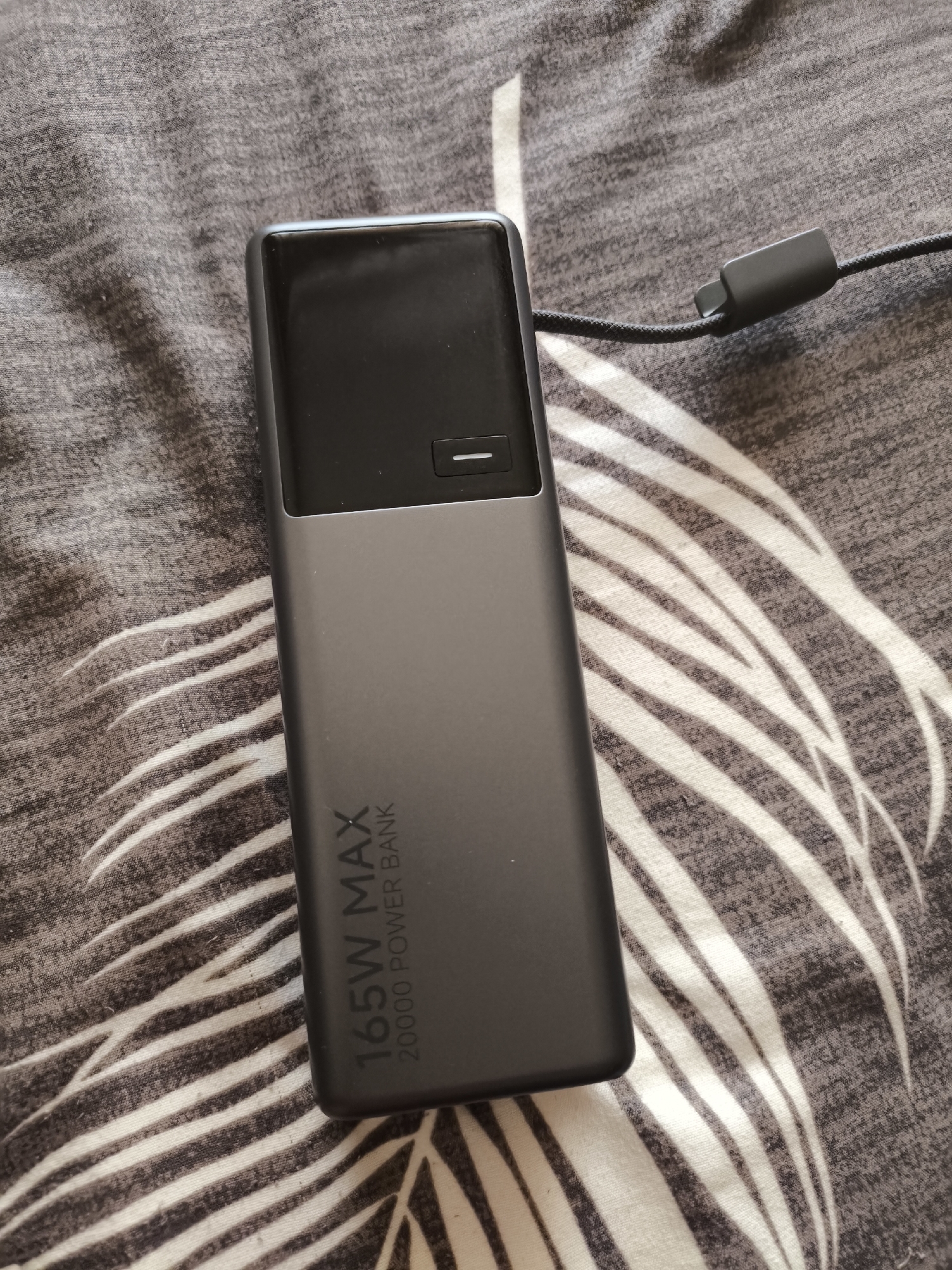 A black 165W MAX 20000 POWER BANK is placed on a grey fabric surface with a white and dark grey striped pattern, likely bedding. The power bank is oriented vertically, with its top end towards the top of the image and its bottom end towards the bottom. The top portion of the power bank is a dark reflective surface, possibly a screen or a glossy finish, with a small horizontal white indicator line. Below this is a matte grey section with the text "165W MAX" and "20000 POWER BANK" printed vertically in white. A black charging cable with a dark grey connector is attached to the power bank and extends out of the frame to the right. The fabric surface appears to be textured and slightly creased. No people are visible in the image. The location in Vsevolozhsk, Russia, is not determinable from the image content.
