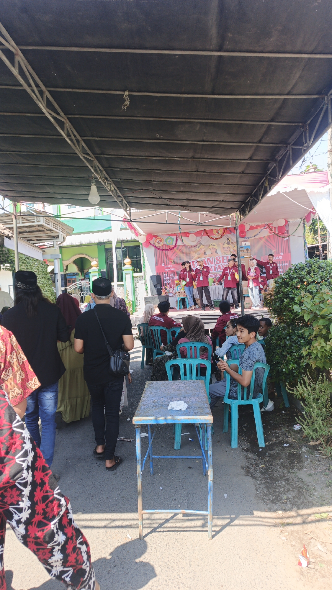 In Suruh, Indonesia, an outdoor stage event is underway. A group of young people in matching maroon shirts and black pants are performing on a stage decorated with balloons and banners that read "EFAN SE" and "AWAR". They appear to be singing or speaking into microphones. Behind them, a building with green and white architecture, possibly a mosque, is visible. A crowd of spectators are seated on blue plastic chairs facing the stage, while others stand around the performance area. Some individuals are wearing traditional Indonesian headwear. A blue foldable table with a white cloth on top sits in the foreground, casting a shadow. The scene is illuminated by bright daylight, suggesting a daytime event. The ground is paved with asphalt, and some areas appear wet or damp. Vegetation, including green bushes, borders the right side of the image.