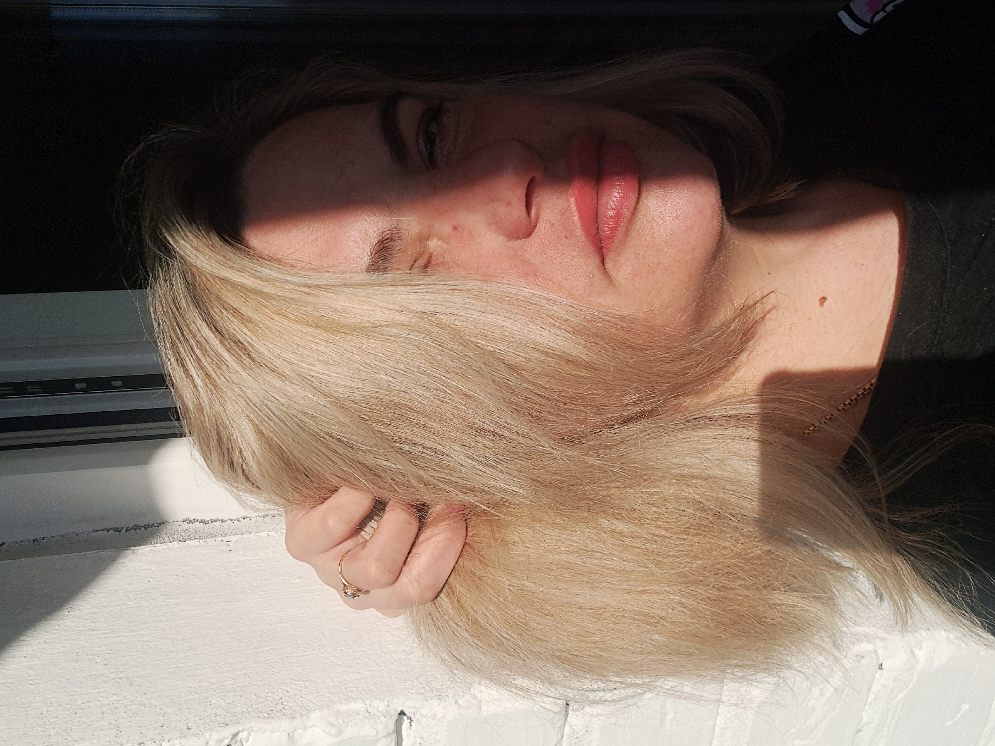 A fair-skinned person with blonde hair is resting their face on a white windowsill. The person is wearing a black shirt, and a gold chain is visible on their neck. Part of their face is illuminated by direct sunlight, while the upper part is in shadow. The person is holding their hair with a ringed hand resting on the windowsill. The background is dark, possibly indicating an indoor setting. Elektrostal, Russia is not confirmed by visual evidence.