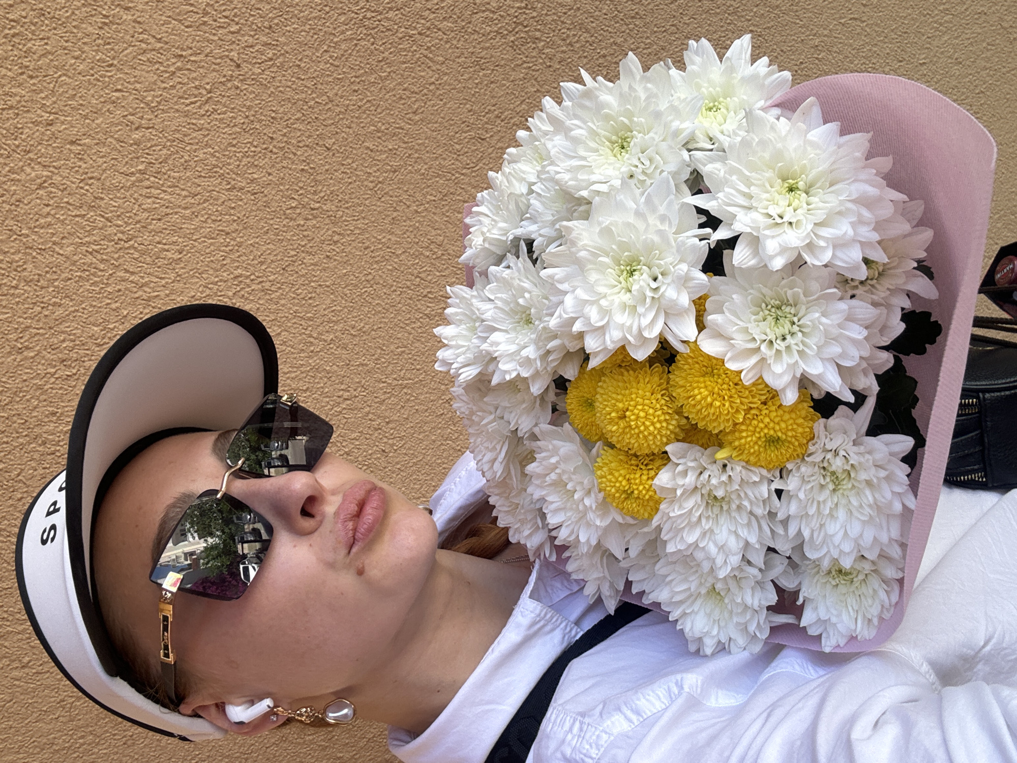A person is positioned against a textured beige wall, possibly outdoors. The individual is wearing a white collared shirt, a black suspender strap, a white visor with black trim and black lettering, sunglasses with a gold chain, and an earpiece with a dangling earring. They are holding a bouquet of white and yellow chrysanthemums wrapped in pink paper. Reflections in the sunglasses show a cityscape with trees. The scene likely takes place during daylight hours in an urban setting but there is nothing indicating this specific location.