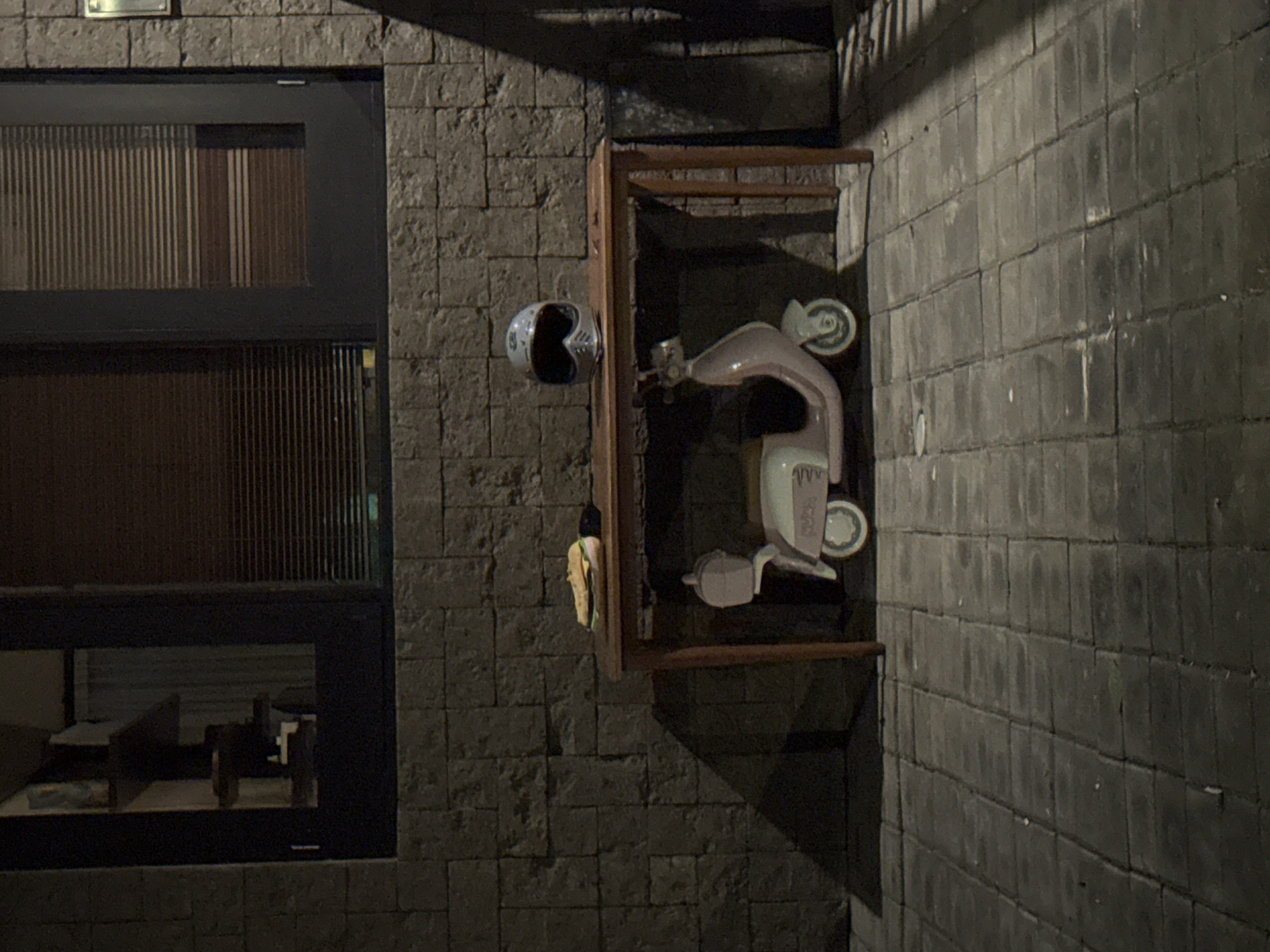 The image shows an outdoor setting in Kuta Selatan, Indonesia, with a dark brick wall. A window with slats and a glass pane is on the left. Attached to the wall is a decorative display containing a vintage-style scooter, a helmet, and a wrapped object. The environment is dimly lit, suggesting it is night. There are no people visible.