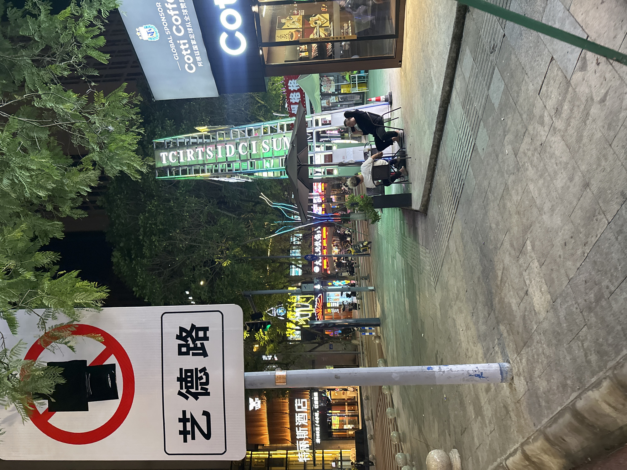 The image depicts a nighttime street scene in the Chongqing High-tech Zone, China. In the foreground, a signpost with a circular red prohibition symbol over a black rectangle is visible, below which is a white rectangle with Chinese characters that read "艺德路" (Yì Dé Lù). The street is paved with stone tiles, and a curb separates the sidewalk from the street.
Further into the scene, two individuals are seated at a small table under a black umbrella, appearing to be patrons of an outdoor seating area. One person is looking at their phone, while the other is engaged in conversation. Behind them, a tall, illuminated structure with green lights and text that appears to say "TCIRTSIDCISUN" rises into the night sky. To the right, a storefront with the "Cotti Coffee" logo is prominently displayed, suggesting a commercial area.
In the background, illuminated buildings with neon signs and streetlights create a vibrant urban atmosphere. Various vehicles, including motorcycles and electric scooters, are parked or in motion on the street, indicating a bustling environment. The overall impression is that of a lively commercial district at night.