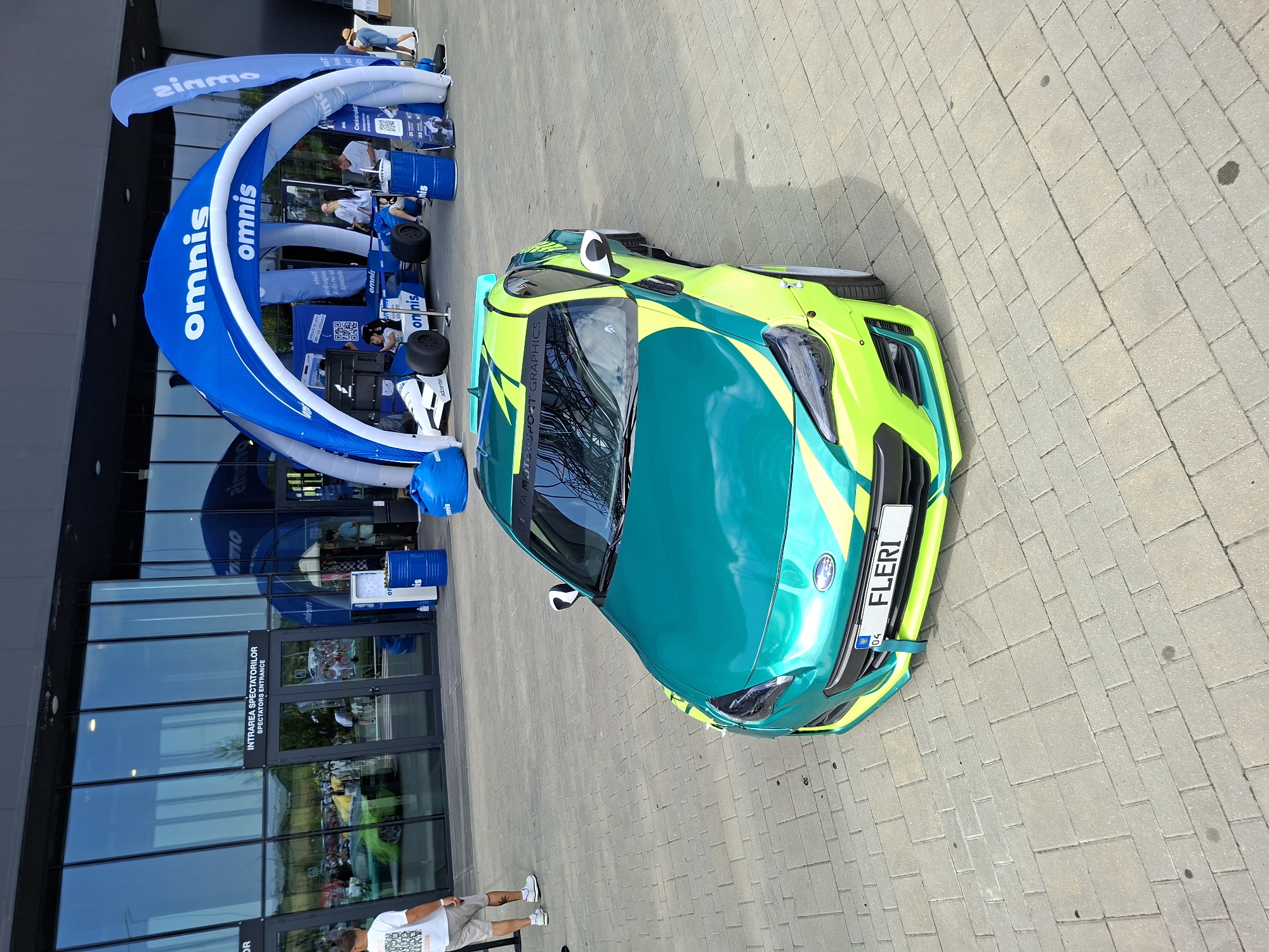 An outdoor event is underway at the Omnis building in Stăuceni, Moldova. In the foreground, a modified Subaru BRZ sports car is parked on a paved area. The car is painted in a striking green and yellow livery, with prominent racing graphics. A license plate with the text "FLERI" is visible.
Behind the car, a large blue inflatable archway with the "omnis" logo spans across the scene, marking an entrance or display area. Underneath the arch, a single-seater race car simulator is set up, with two children seated inside. Several people are milling around the event, some standing and observing, while others appear to be engaged in activities. The Omnis building, with its large glass facade, serves as the backdrop, reflecting the surrounding environment and the event's activities. The overall scene suggests a car show or a promotional event.