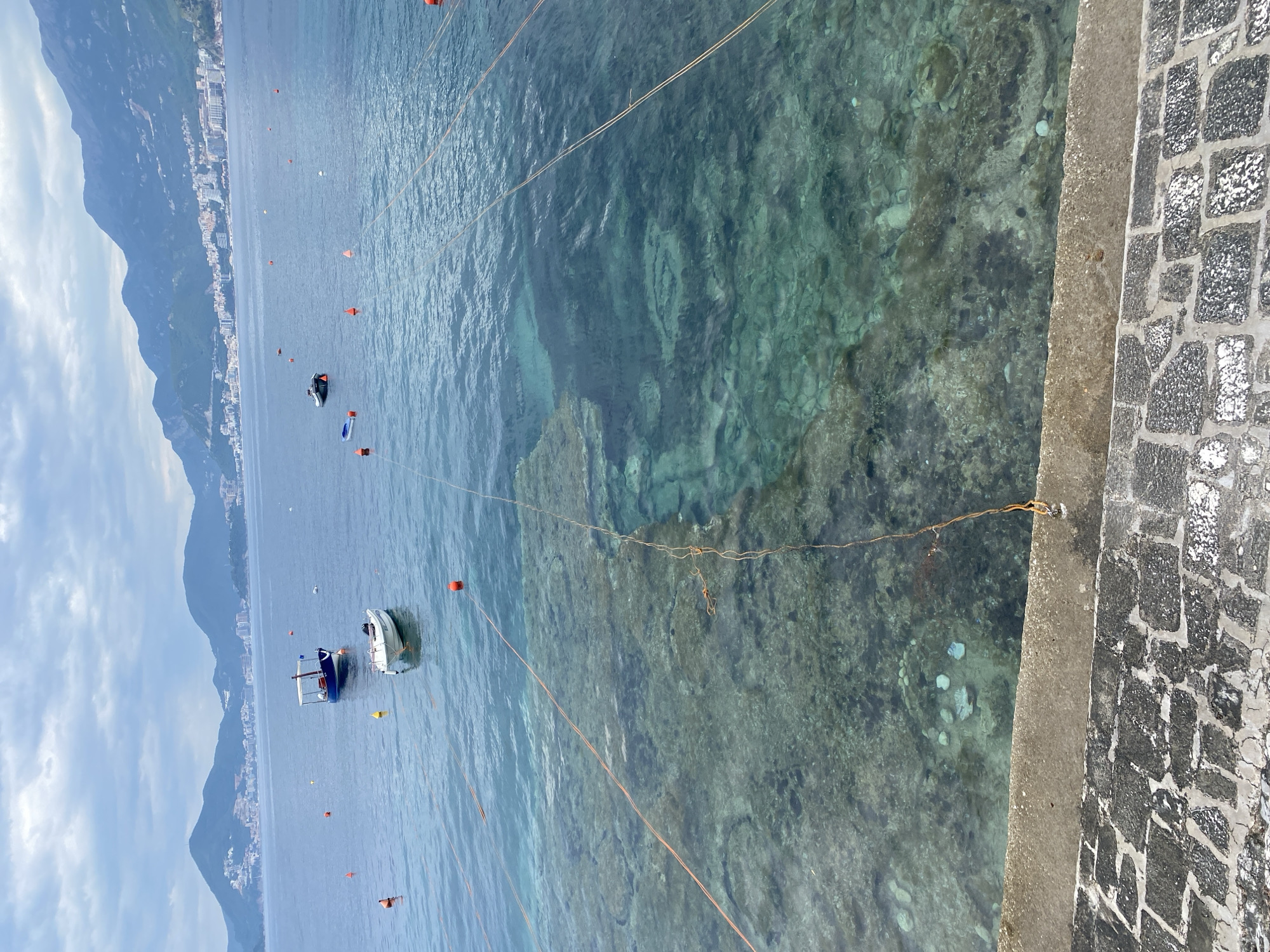 From a high angle, a view captures the cityscape of Sveti Stefan, Montenegro, from an elevated position. The scene includes the Adriatic Sea, where several small boats are anchored. The sea's transparency reveals the submerged rocky terrain below the water's surface. A stone-paved area is visible on the right edge of the image. Overhead, lines or ropes are stretched across the scene. The sky above is partly cloudy.